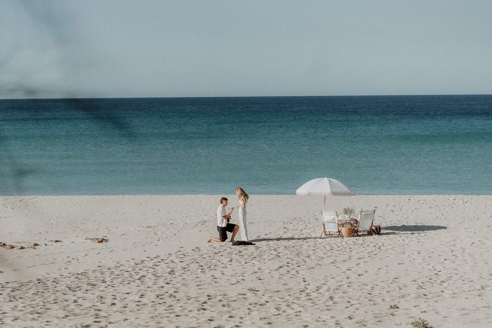 A couple happily getting engaged at Eagle Bay Beach, surrounded by a luxurious white picnic setup including a fringed umbrella, deck chairs, and rattan coffee table.