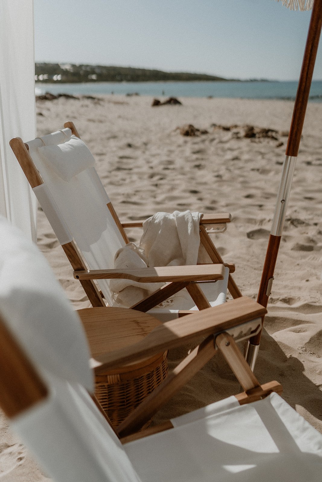 A couple happily getting engaged at Eagle Bay Beach, surrounded by a luxurious white picnic setup including a fringed cabana, deck chairs, and rattan coffee table.