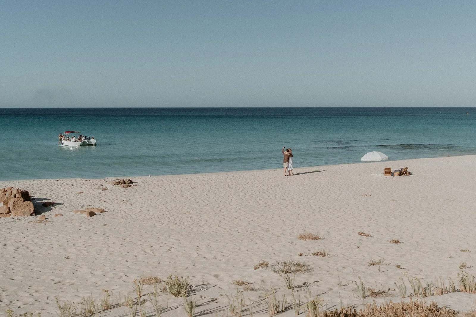 A couple happily getting engaged at Eagle Bay Beach, surrounded by a luxurious white picnic setup including a fringed umbrella, deck chairs, and rattan coffee table.