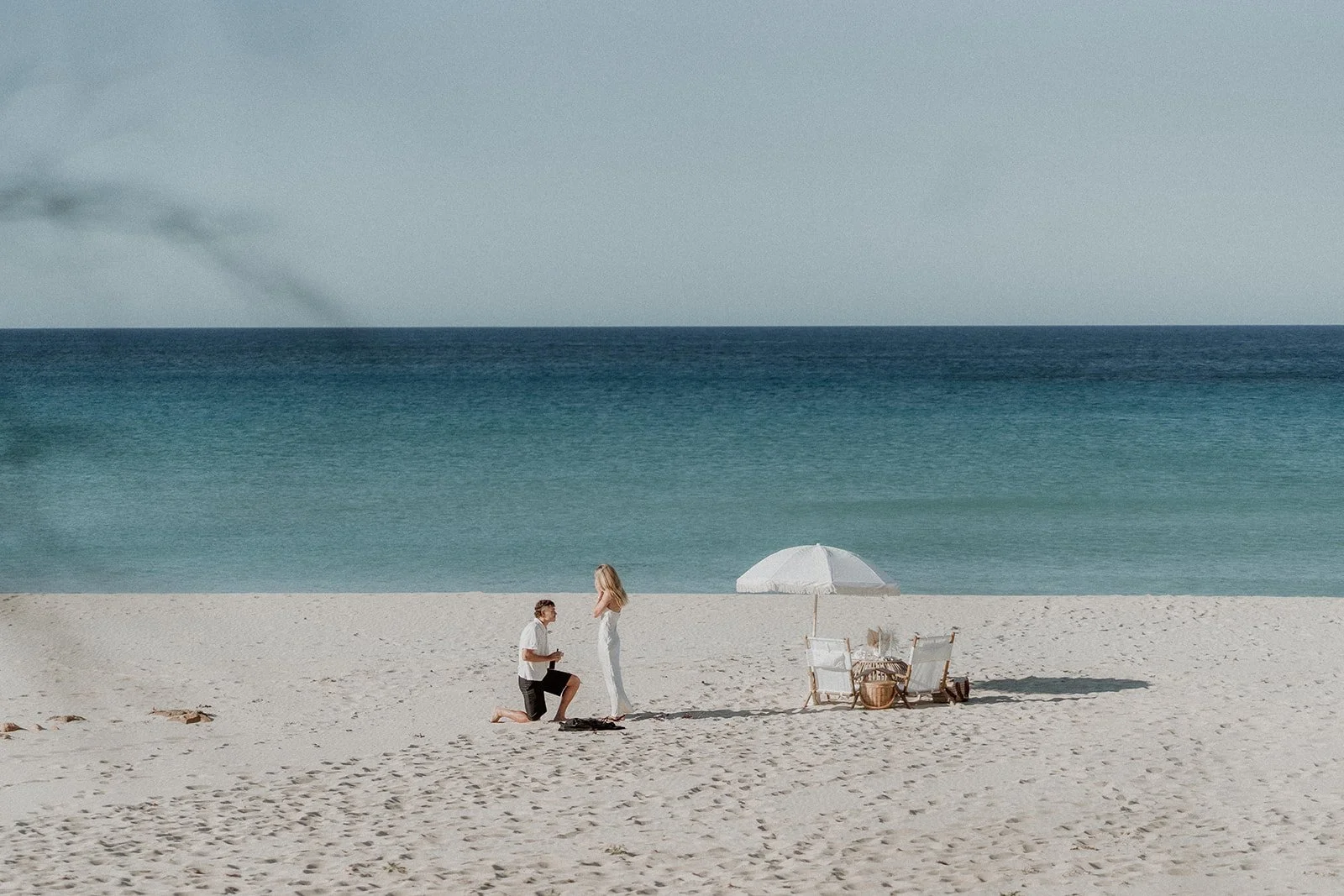A couple happily getting engaged at Eagle Bay Beach, surrounded by a luxurious white picnic setup including a fringed umbrella, deck chairs, and rattan coffee table.