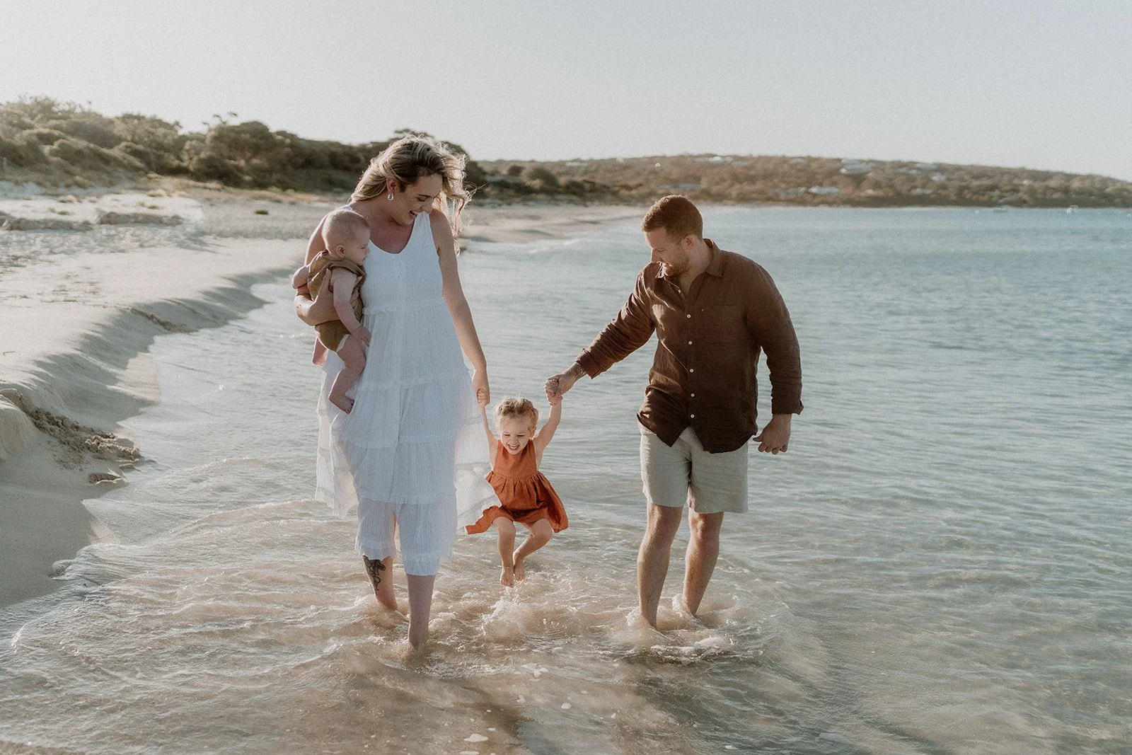 A couple happily getting engaged at Eagle Bay Beach, surrounded by a luxurious white picnic setup including a fringed umbrella, deck chairs, and rattan coffee table.