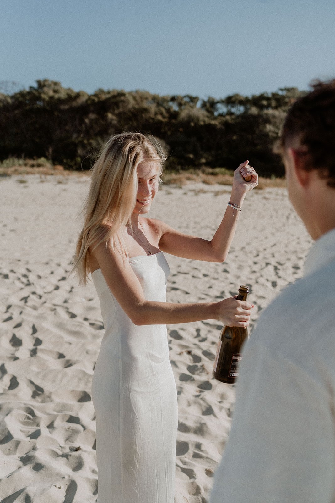A couple happily getting engaged at Eagle Bay Beach, surrounded by a luxurious white picnic setup including a fringed umbrella, deck chairs, and rattan coffee table.