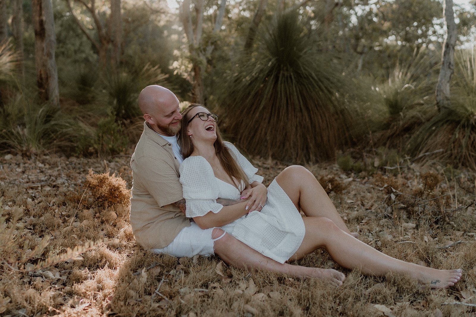A couple happily getting engaged at Eagle Bay Beach, surrounded by a luxurious white picnic setup including a fringed cabana, deck chairs, and rattan coffee table.