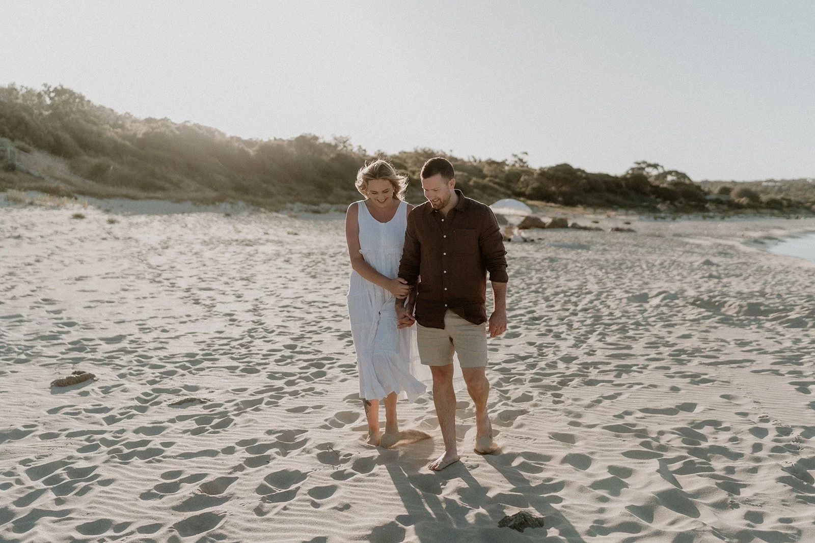 A couple happily getting engaged at Eagle Bay Beach, surrounded by a luxurious white picnic setup including a fringed umbrella, deck chairs, and rattan coffee table.