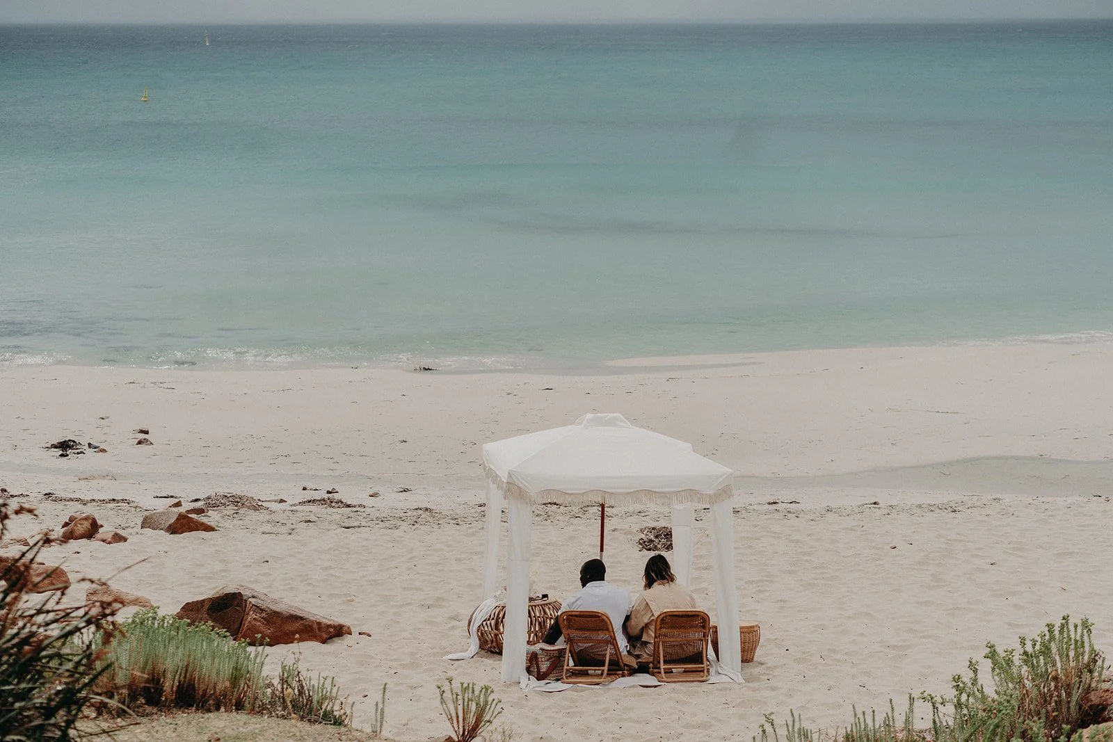A couple happily getting engaged at Meelup Beach, surrounded by a luxurious white picnic setup including a fringed cabana, deck chairs, and rattan coffee table.