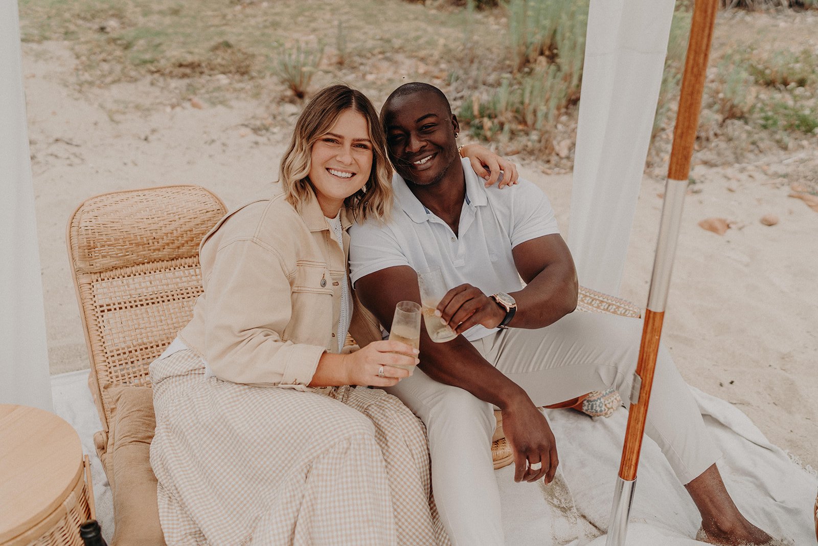 A couple happily getting engaged at Meelup Beach, surrounded by a luxurious white picnic setup including a fringed cabana, deck chairs, and rattan coffee table.