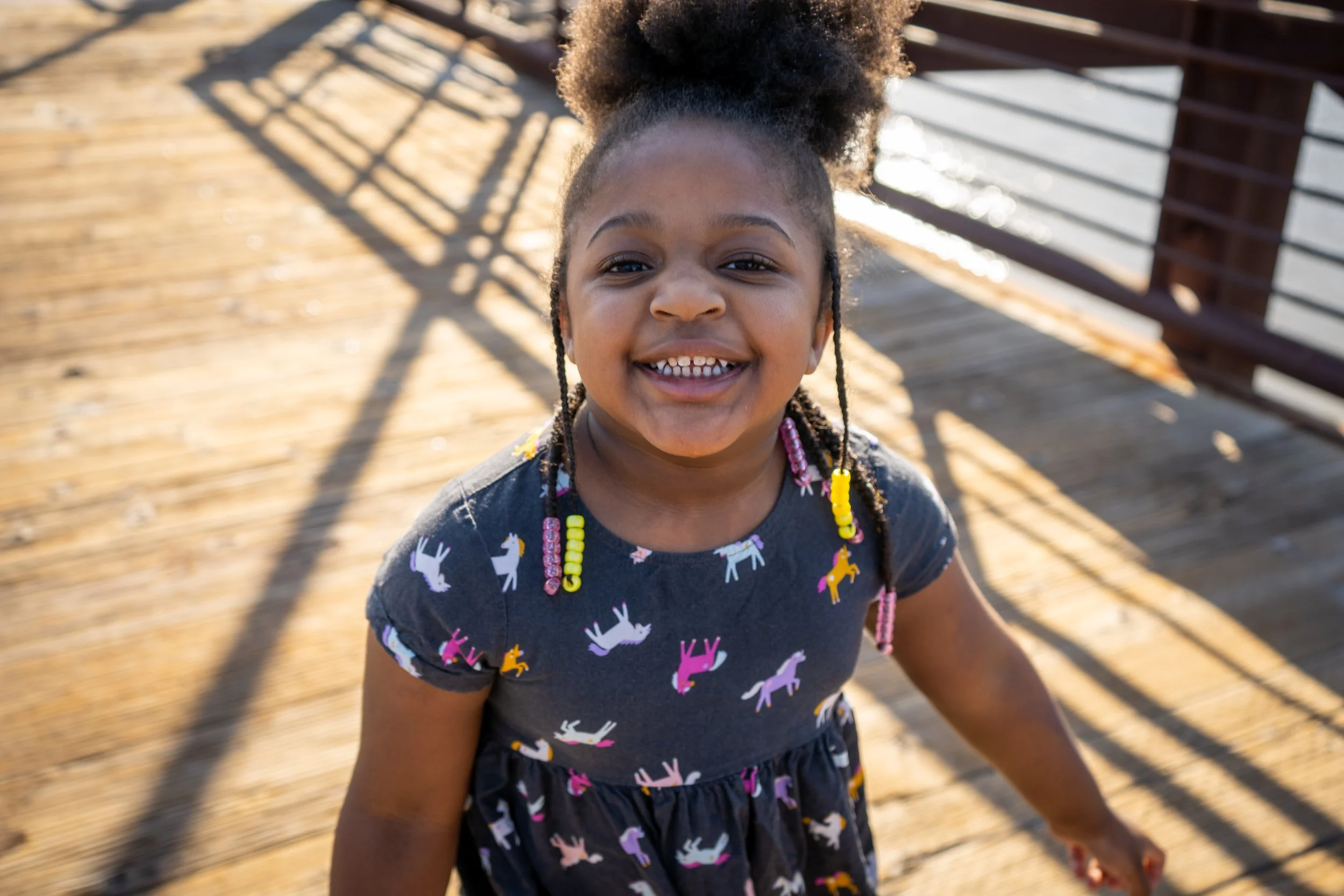A LITTLE GIRL STANDING ON A BRIDGE LOOKS AT THE CAMERA WITH A MISSING FRONT TOOTH
