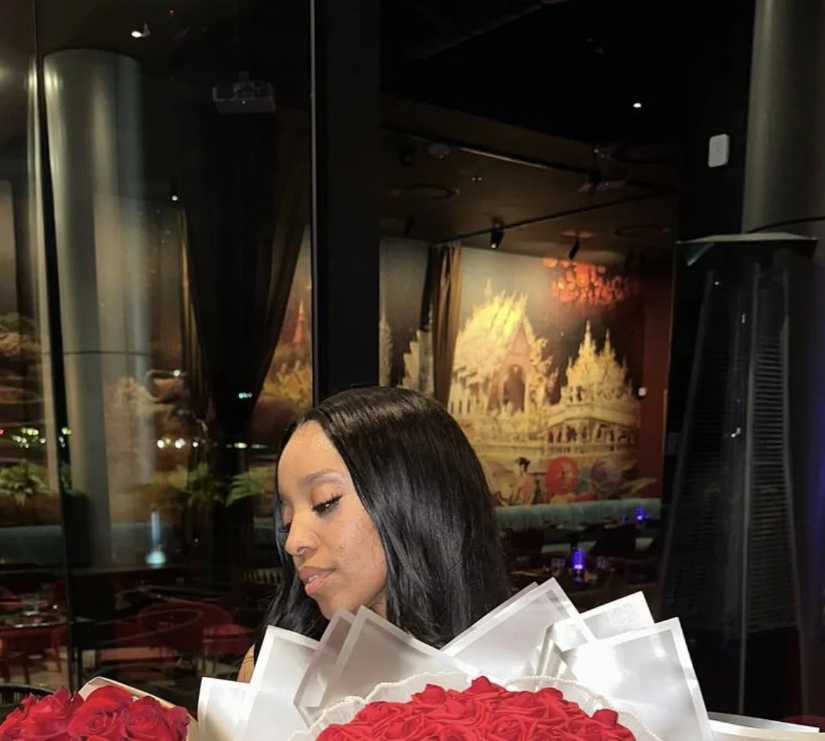 A woman with long black hair is sitting in a dimly lit restaurant, surrounded by a bouquet of red roses, with a decorative Asian-themed mural in the background.