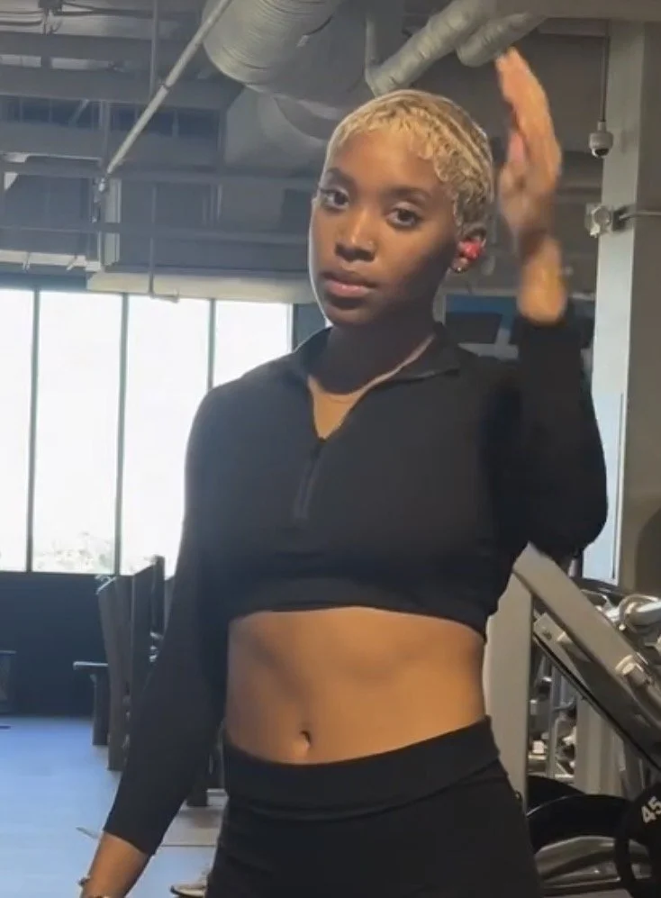 A woman standing in a gym, wiping sweat from her face with a towel, wearing a black crop top and leggings.