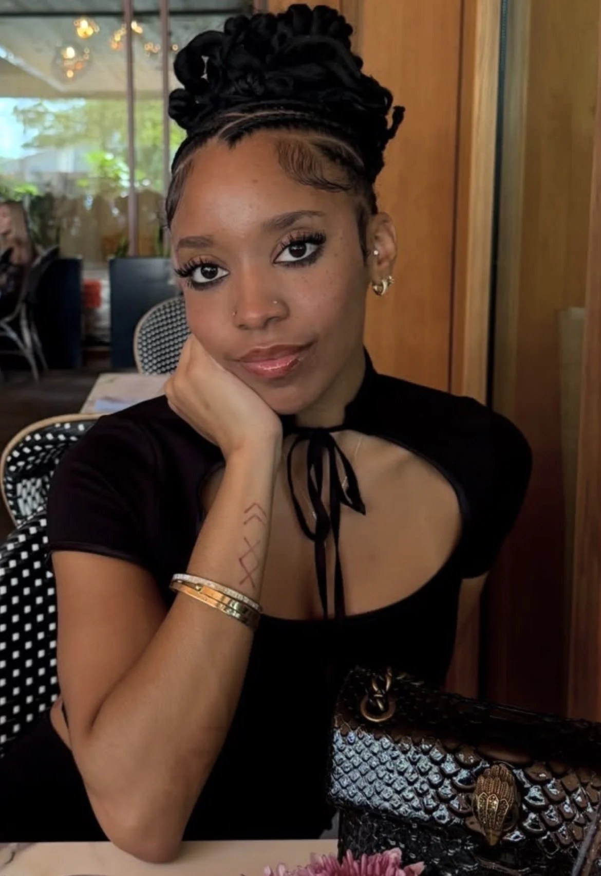 A young woman with braided hair styled in an updo, wearing a black top with a tie at the neckline, gold bracelets, and earrings. She is sitting at a table with her chin resting on her hand, looking at the camera in a restaurant or cafe setting.