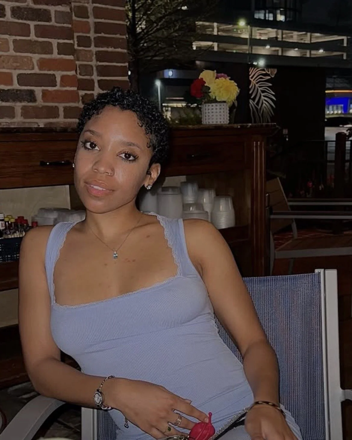 A young woman with short curly black hair, wearing a light gray tank top with lace trim, sitting at a restaurant or cafe at night. She has jewelry including earrings, a necklace, and bracelets, and is holding a drink with a red umbrella. The background features a brick wall, a wooden shelf with plates, and a vase of colorful flowers, with a view of a multi-story building and street lights outside.