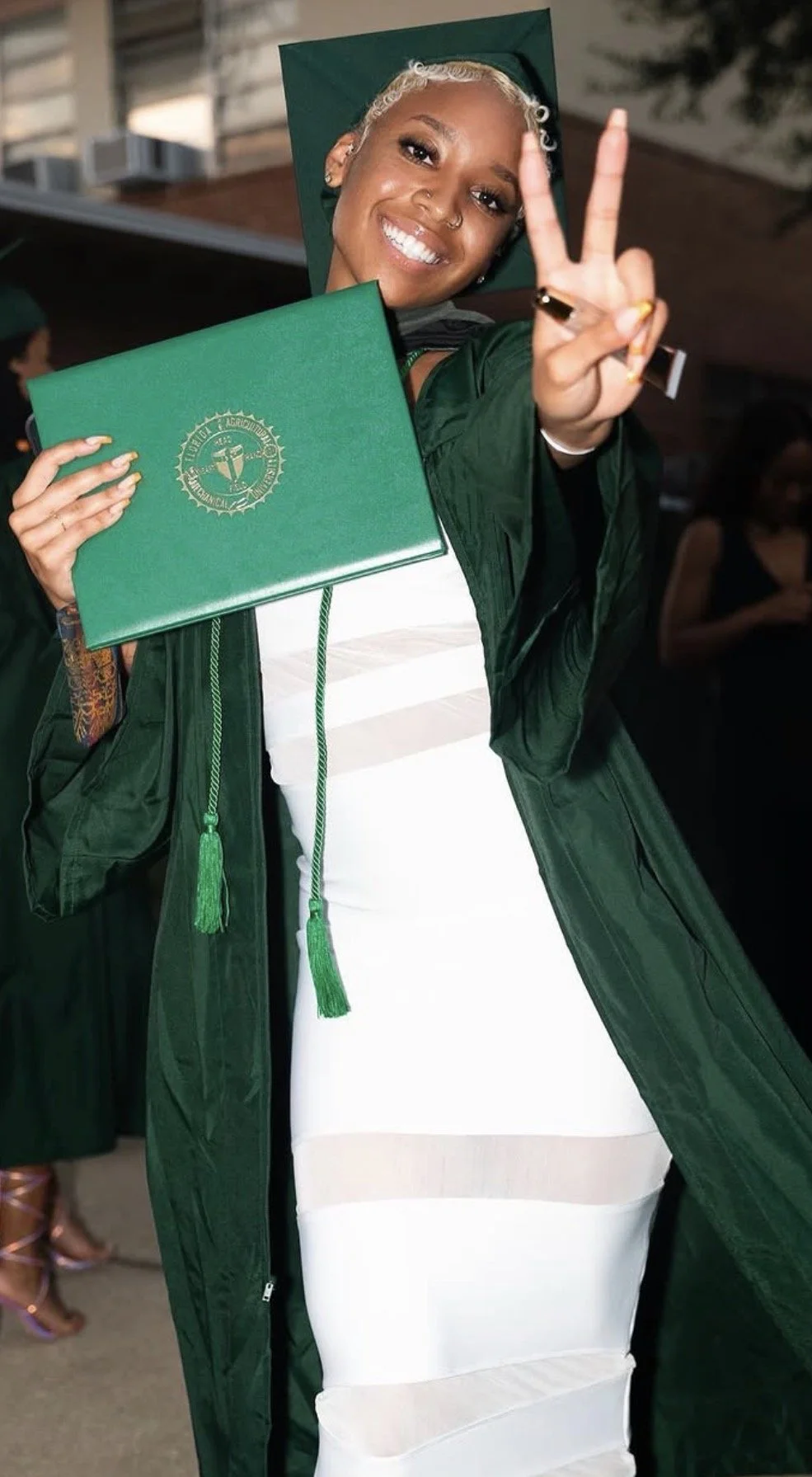A young woman in a graduation cap and gown holding her diploma and flashing a peace sign, smiling for the camera.
