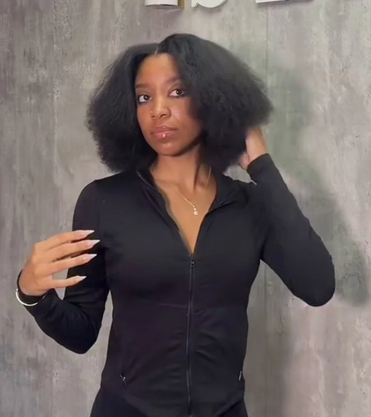 A woman with voluminous black natural hair wearing a black zip-up jacket, standing against a gray textured wall.