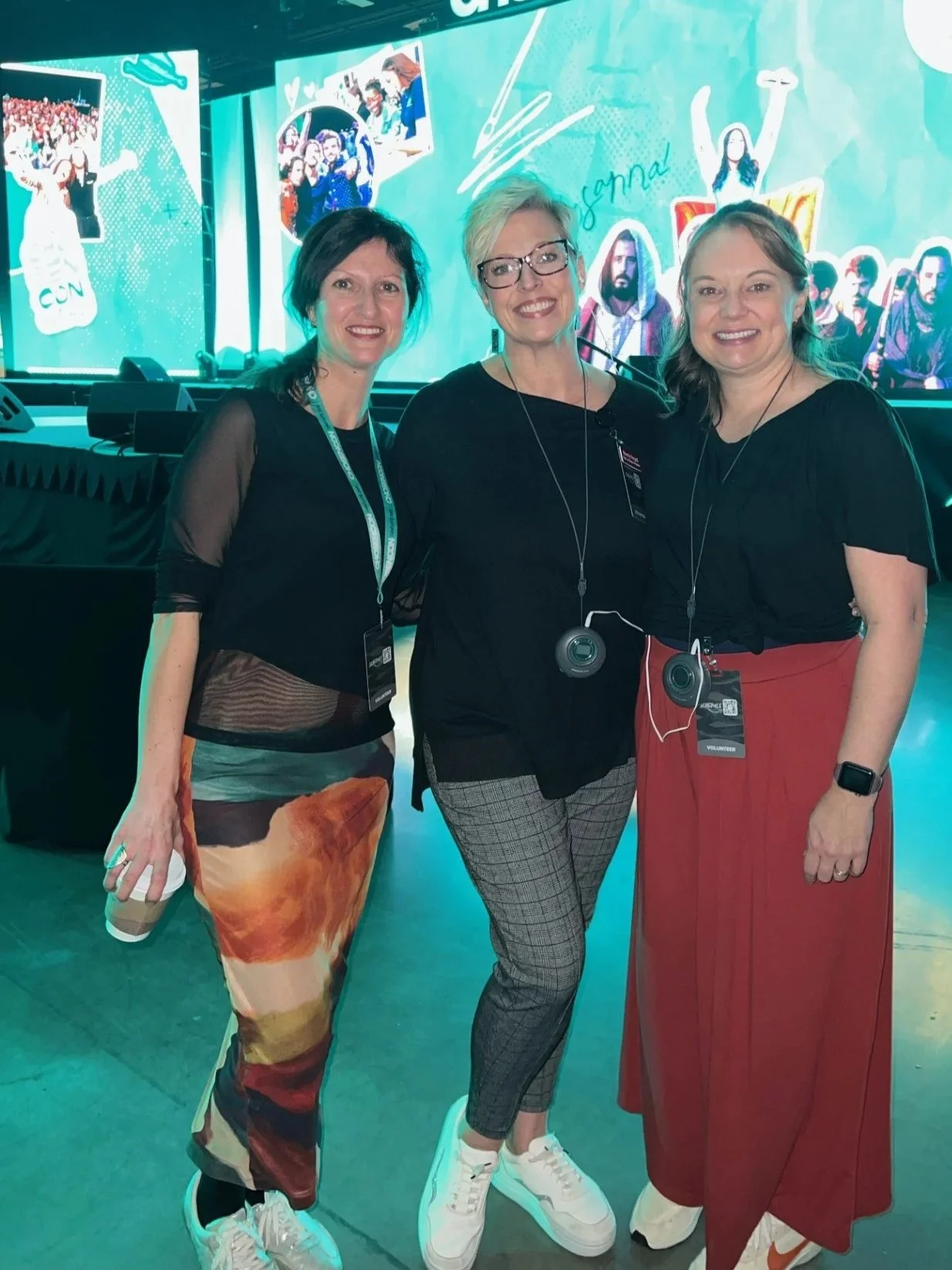 Three women standing together at an event or conference with a large screen in the background displaying an animated scene with people and graphics.