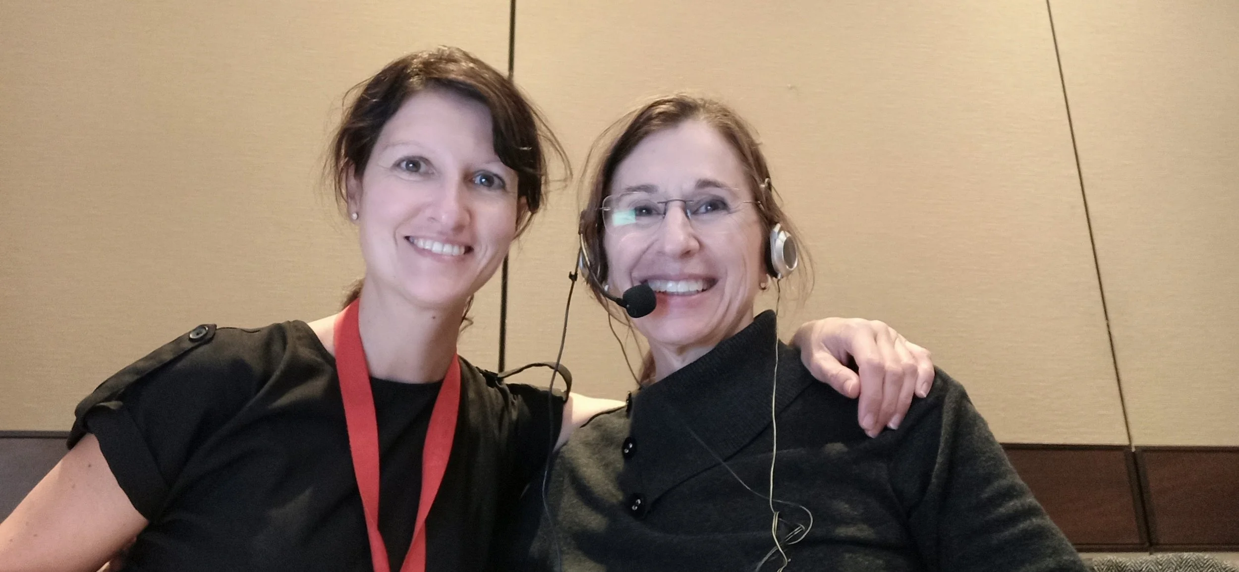 Two women smiling, one wearing a black top and a red lanyard, the other wearing glasses, a gray top, and a headset, posing together with their arms around each other against a beige wall.