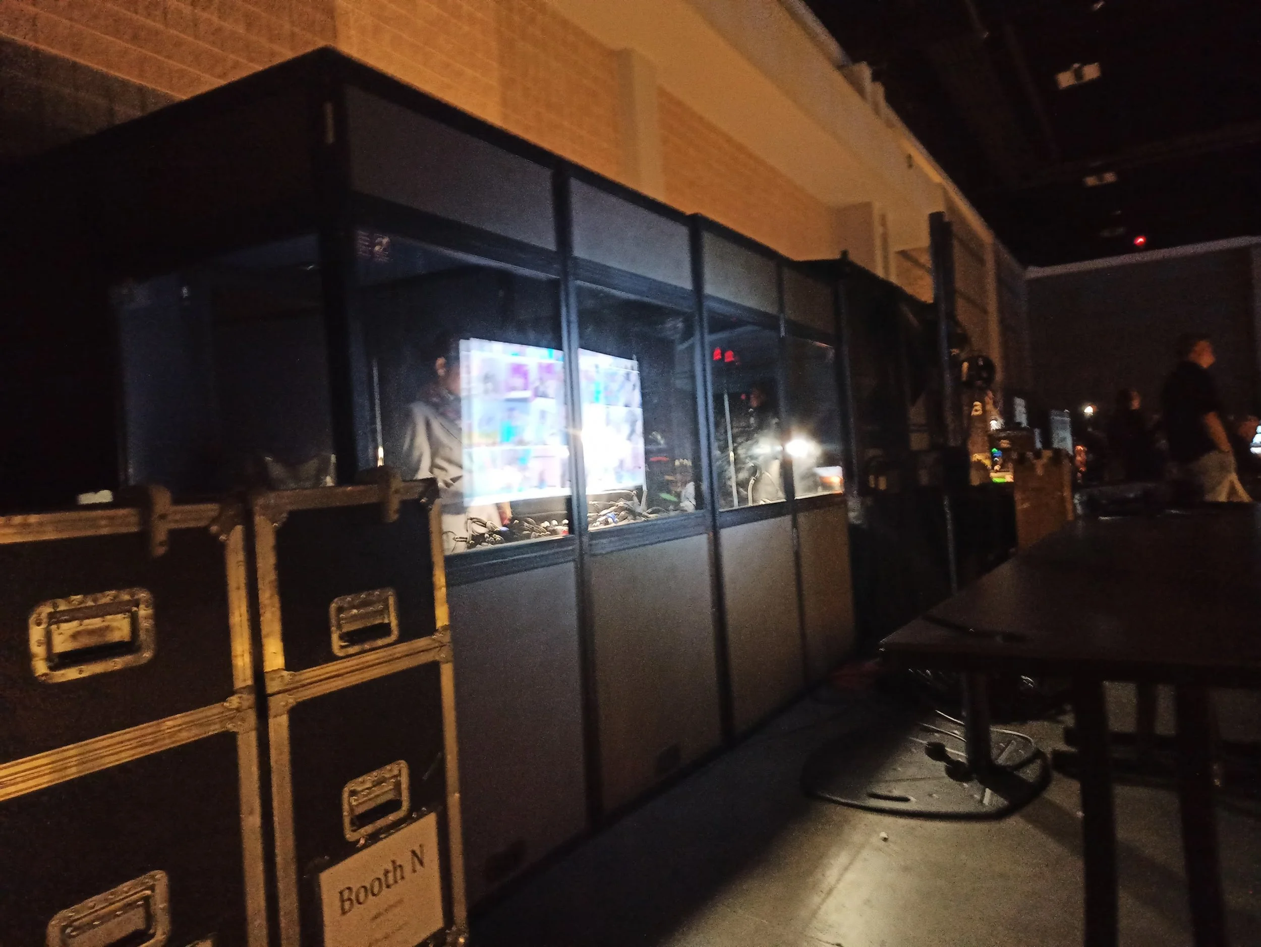 A dark indoor venue with DJ equipment and booth, with a person visible inside behind a glass window, several black storage cases labeled 'Booth N', and people in the background.