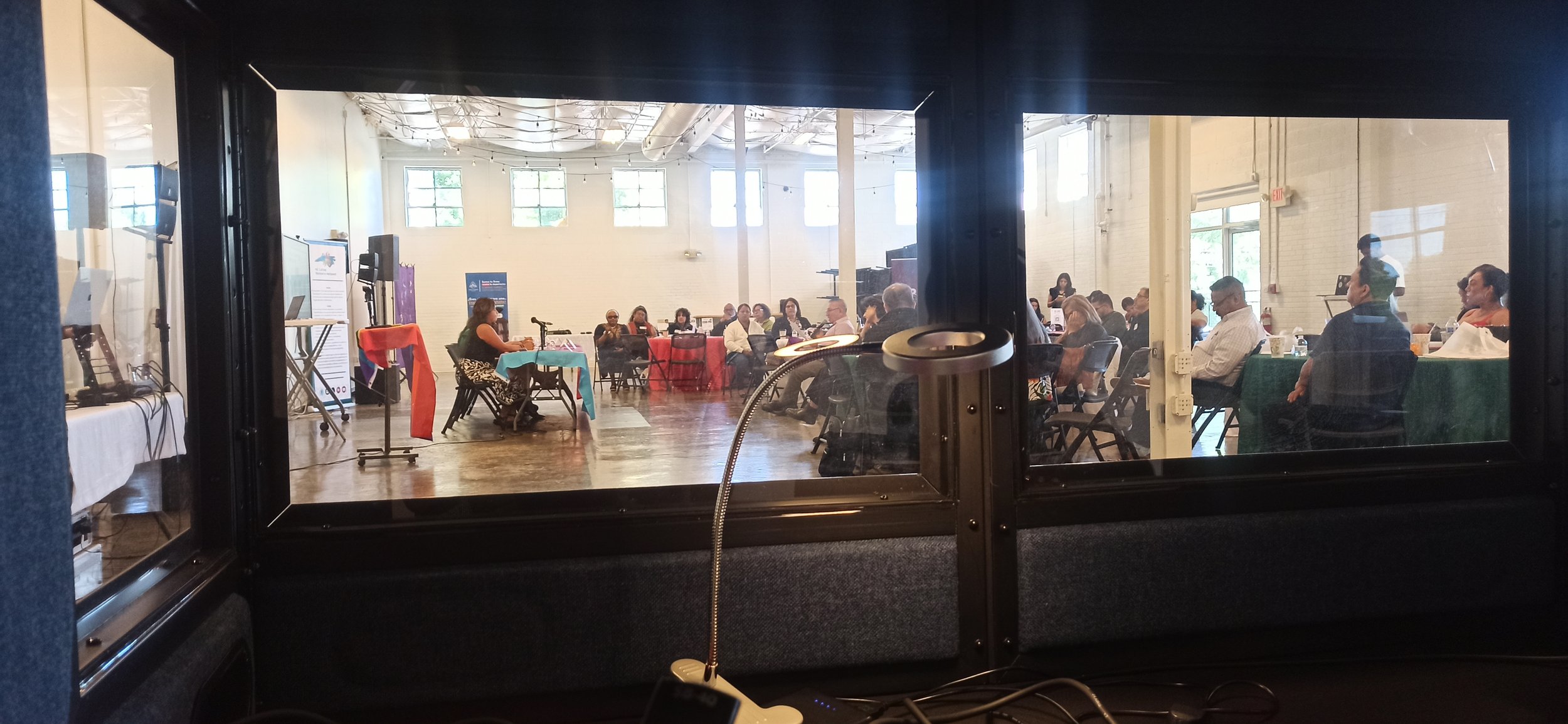 View of a large room with people seated at tables, seen through a glass window from behind a desk with a lamp and some equipment.
