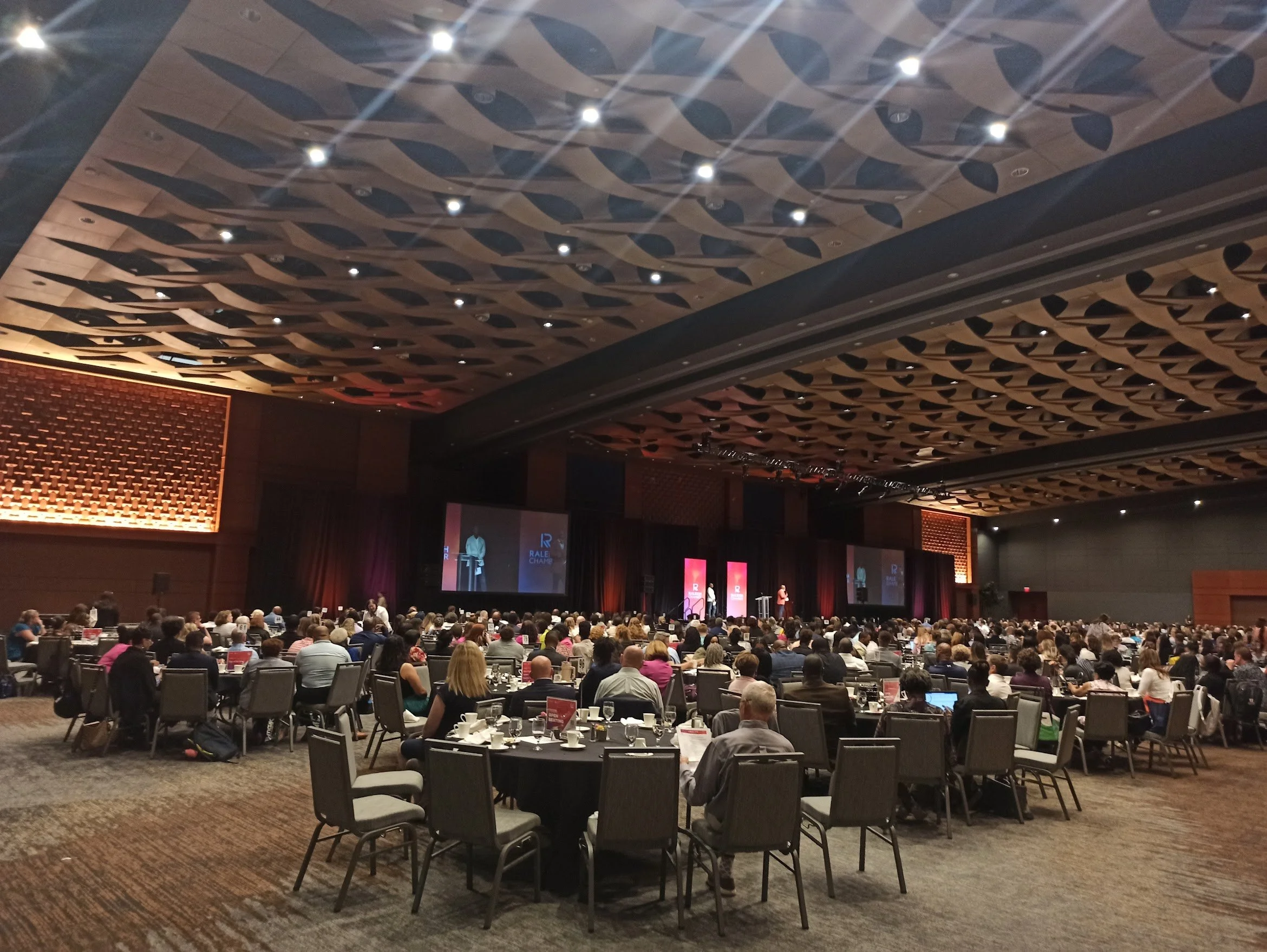 Large conference room filled with round tables of attendees listening to a speaker on stage, with multiple screens displaying the presentation.