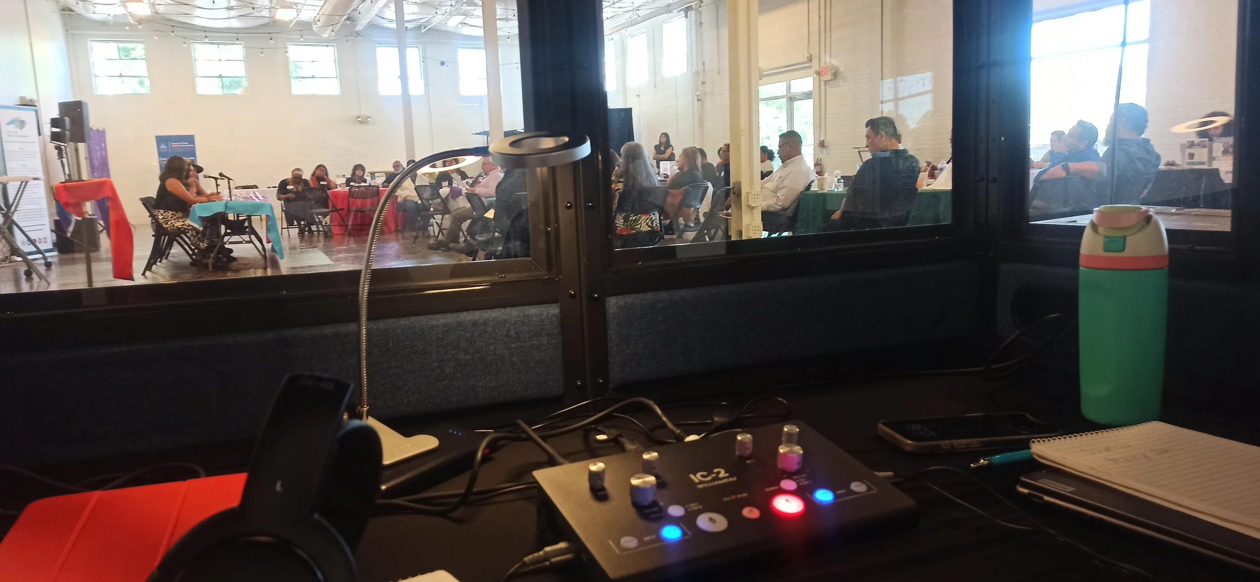 View from behind a sound or DJ booth looking into a conference or seminar room with people seated at tables, some using laptops and listening to a speaker at a podium.