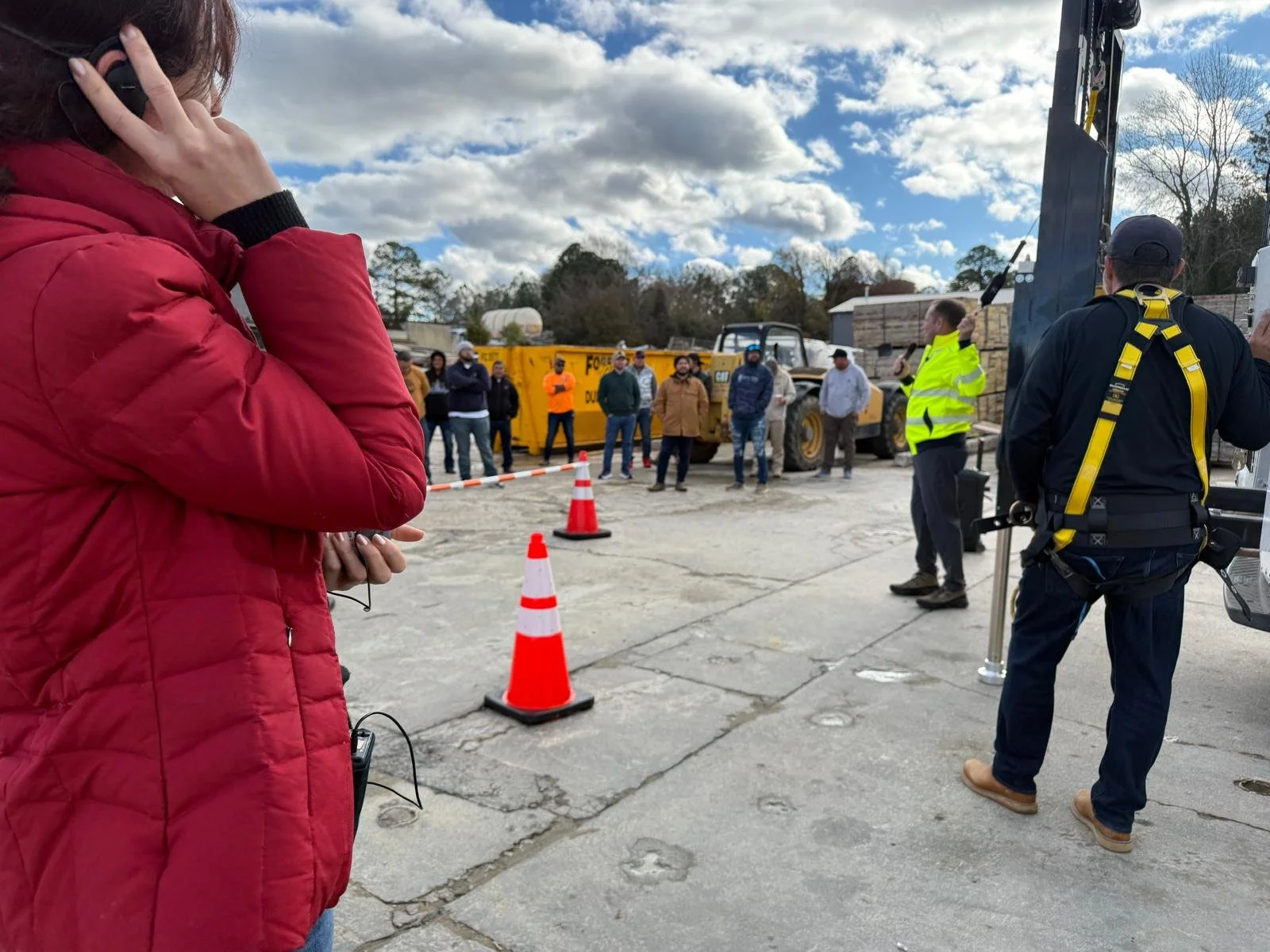 A group of construction workers and officials gathered outdoors, with some wearing safety vests, standing near construction equipment and orange cones on a cloudy day.