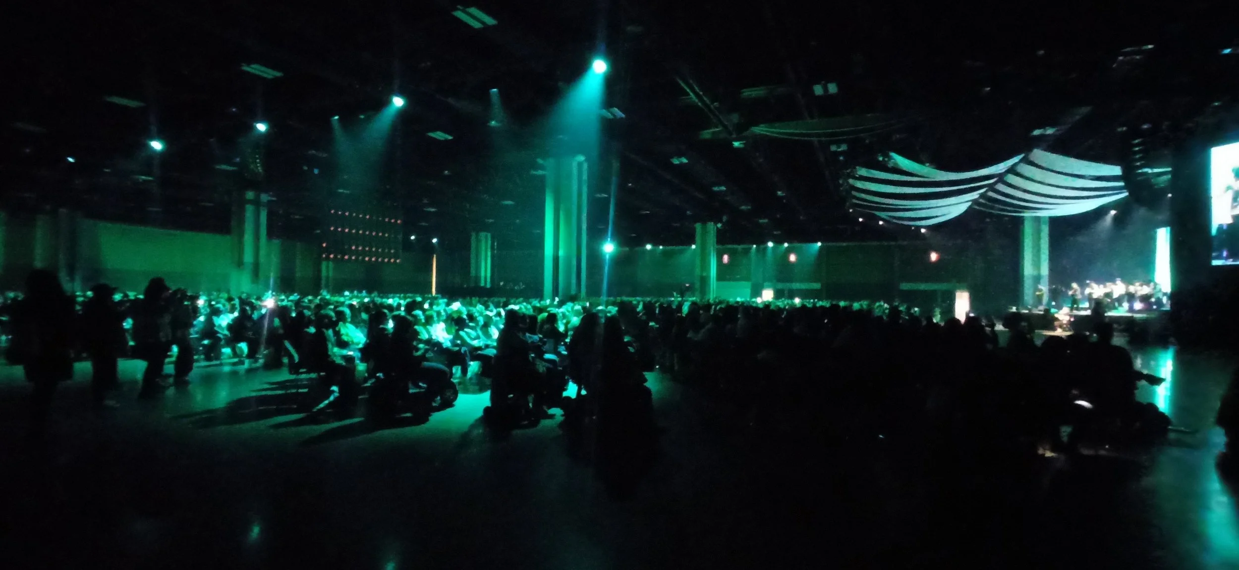 Crowd of people seated in a dark, green-lit auditorium or conference hall attending a presentation or performance.