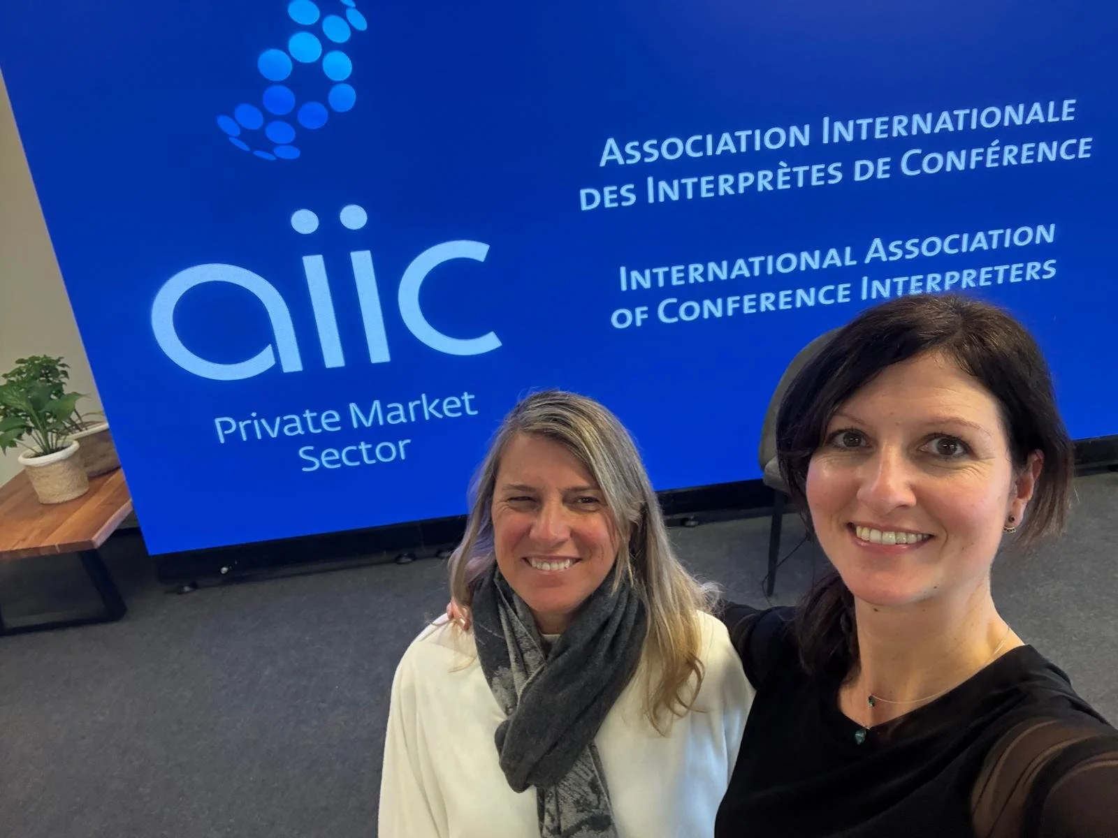 Two women smiling for a selfie in front of a blue sign that reads 'International Association of Conference Interpreters,' 'Private Market Sector,' with a logo at the top left.