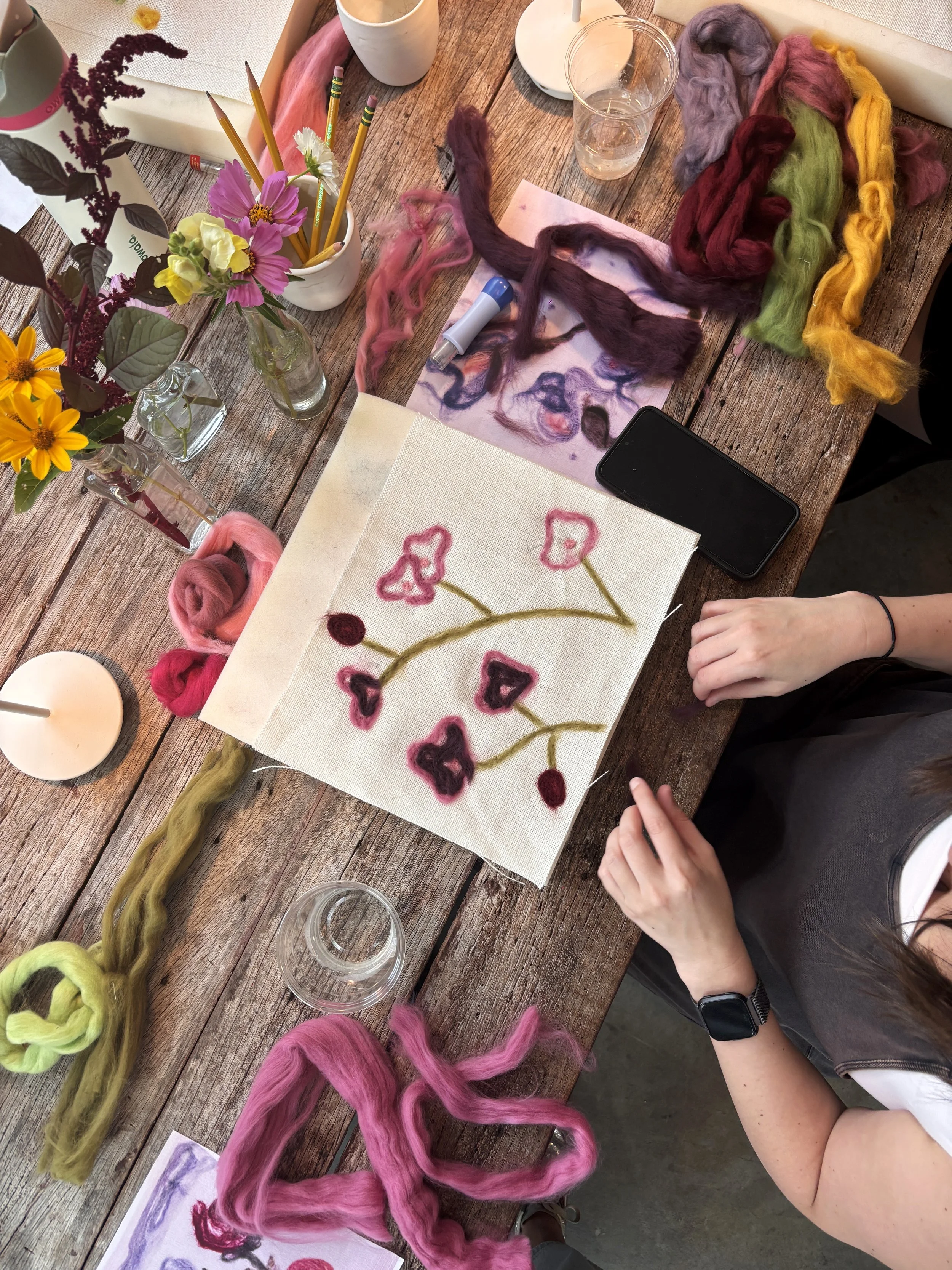 Guest creating a Paint with Wool piece, Le Studio Anthost, New York, with wool roving, floral arrangements, and art supplies on the table.