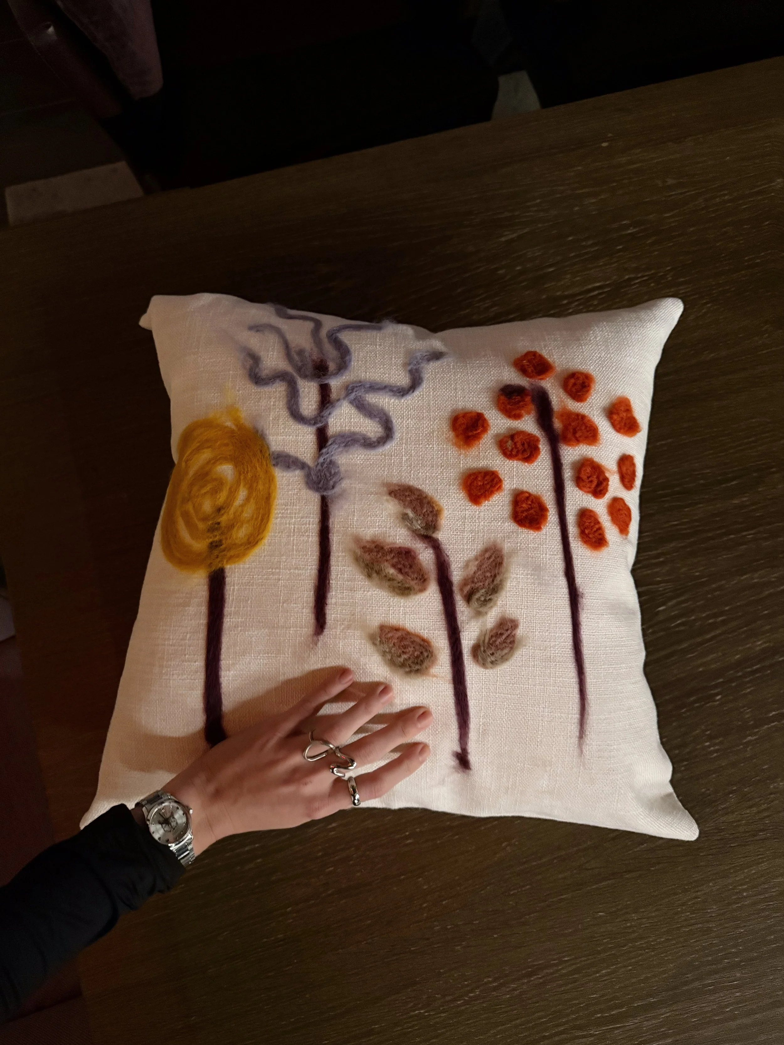 A close-up view of a decorative pillow with embroidered abstract trees in various colors on a wooden surface, with a person's hand and wristwatch visible in the lower left corner.