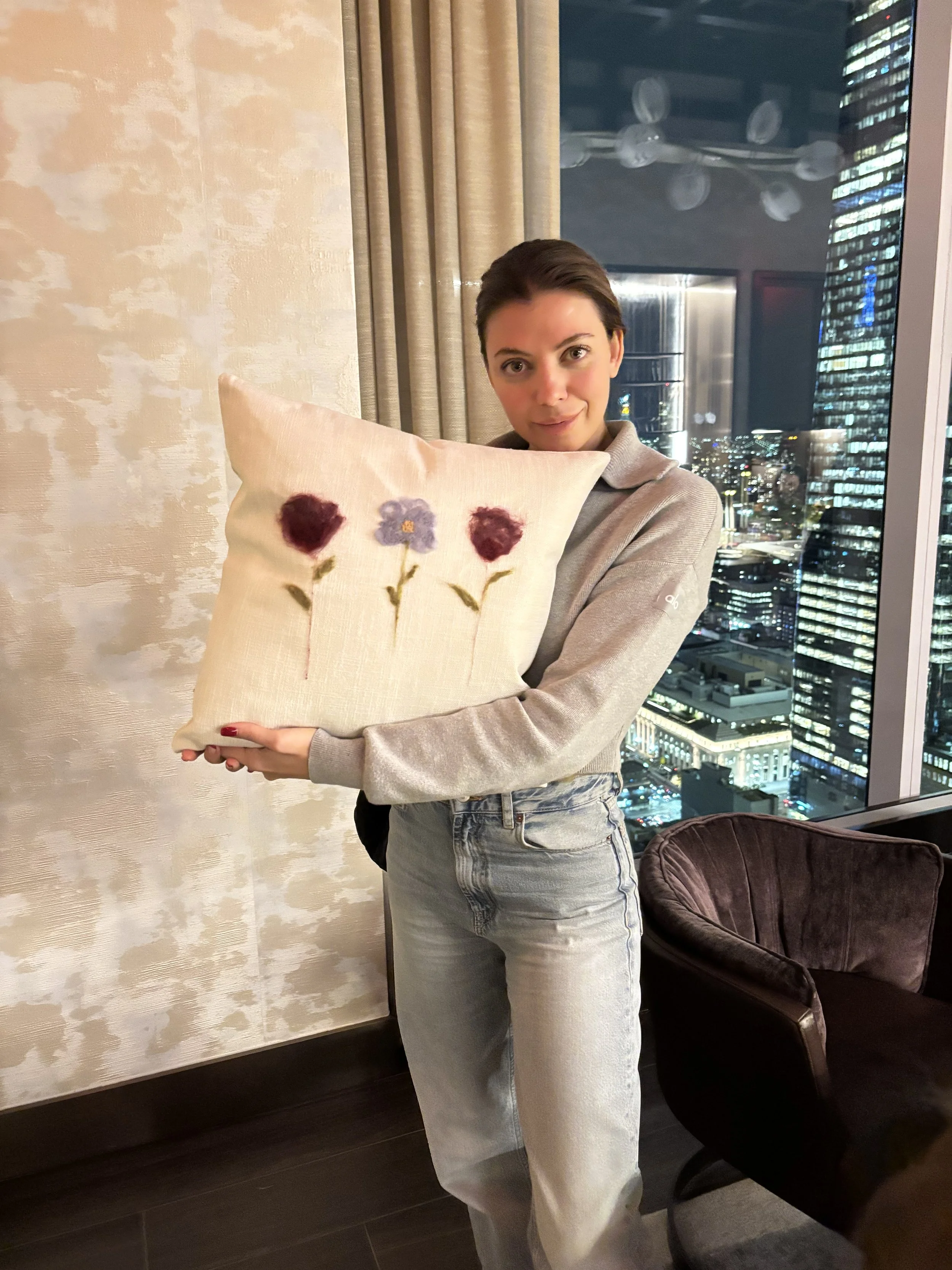 A woman with short dark hair holding a beige pillow with purple flower embroidery in front of her. She is standing in a room with city lights and tall buildings visible outside a window.