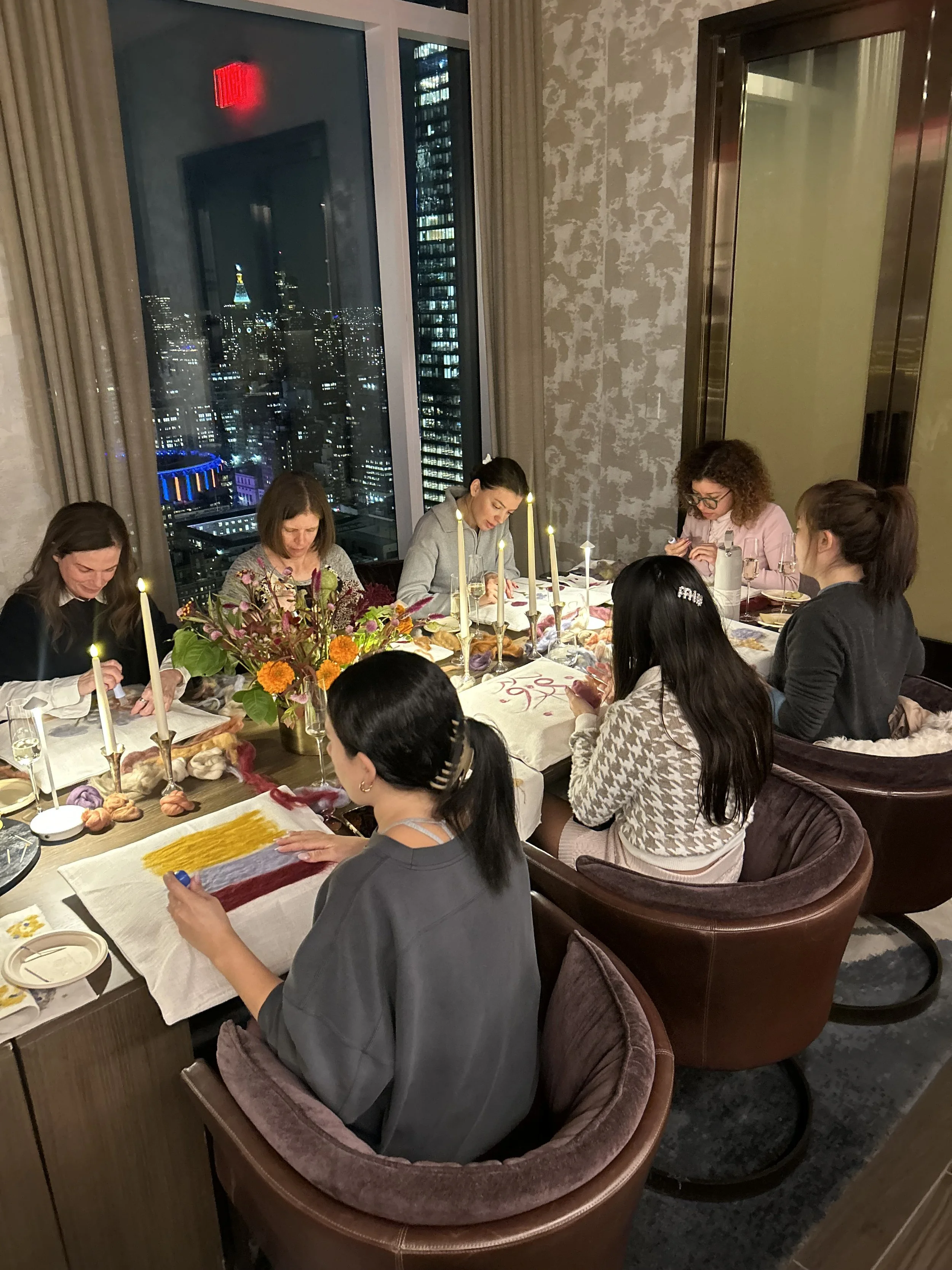 People sitting around a dinner table in a high-rise apartment at night, with city lights and a tall building with colorful lights visible through large windows.