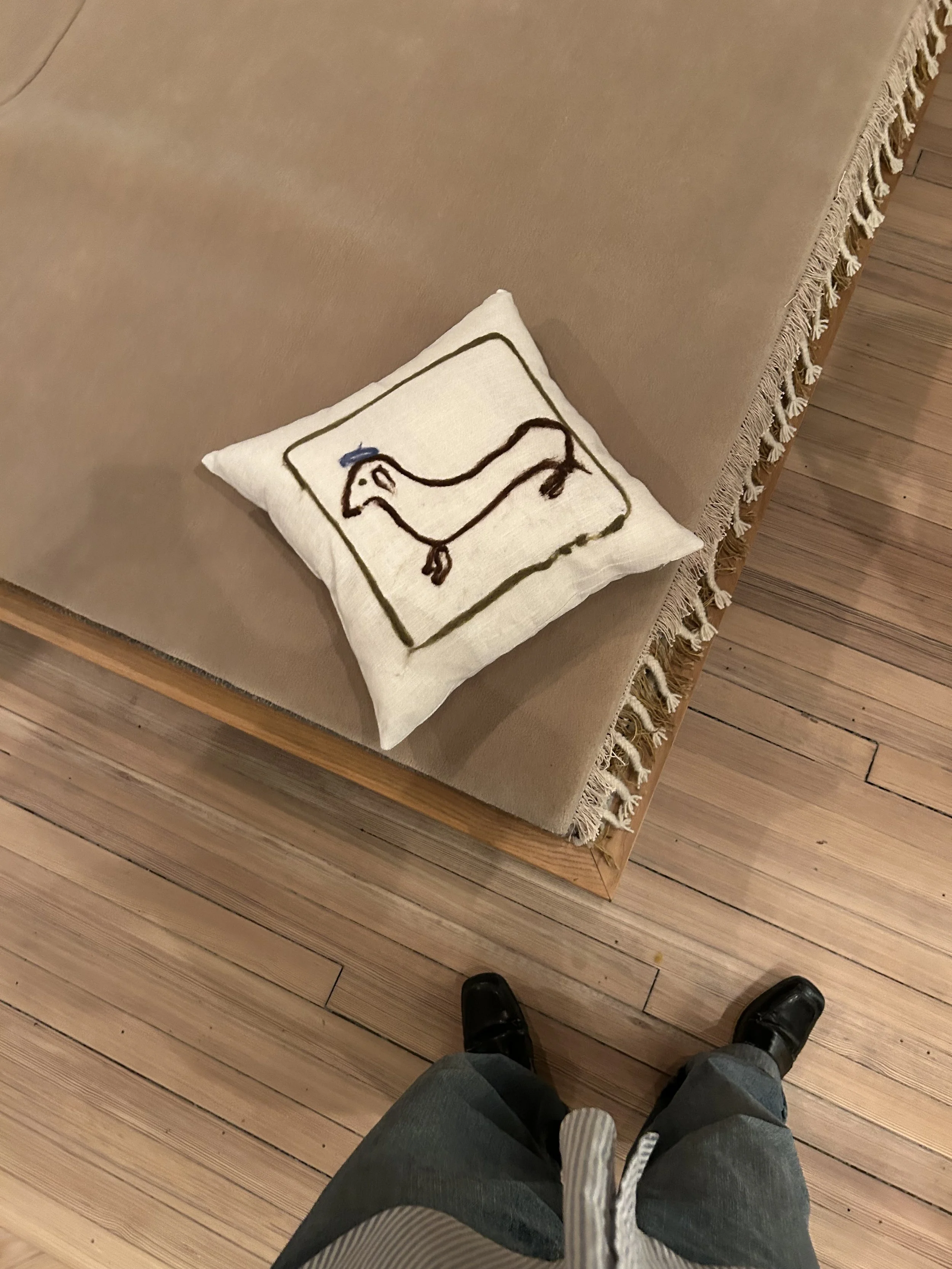 A person standing on a wooden floor wearing black shoes and gray pants, looking down at a beige cushioned bench with a pillow on top. The pillow has a drawing of a bird within a rectangular border.