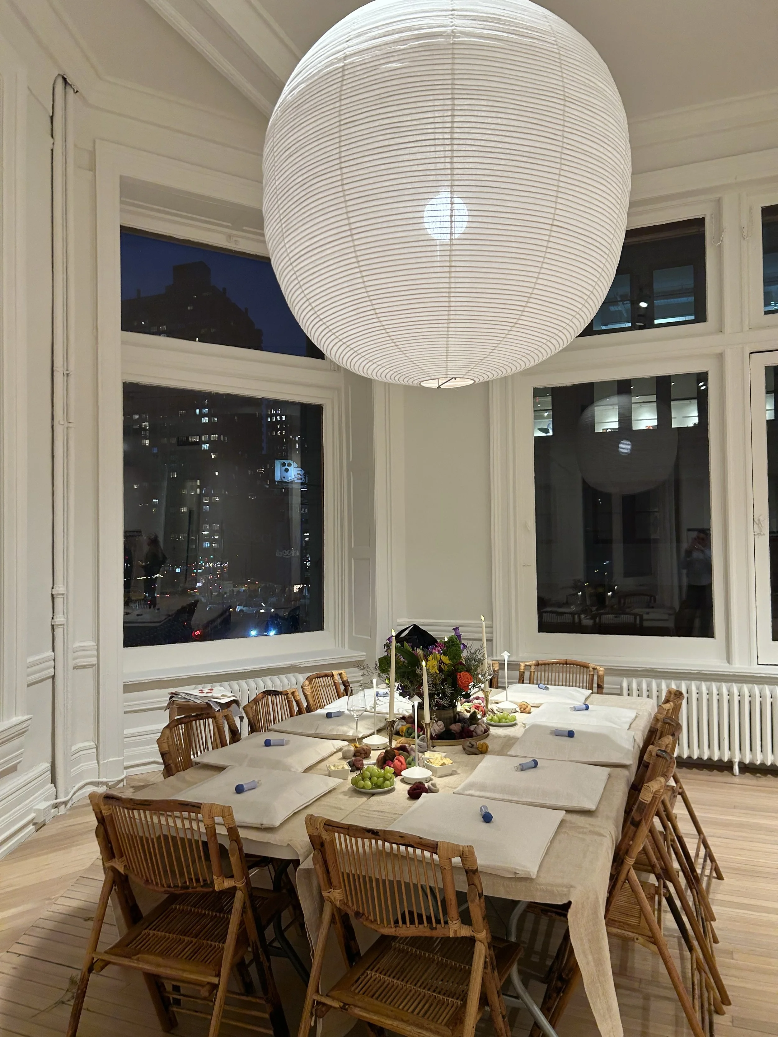A dining room with a long table set for a celebration, with a floral centerpiece and several plates of food and grapes, surrounded by wooden chairs. Large windows show a cityscape at night, and a large, white paper lantern hangs from the ceiling illuminating the room.