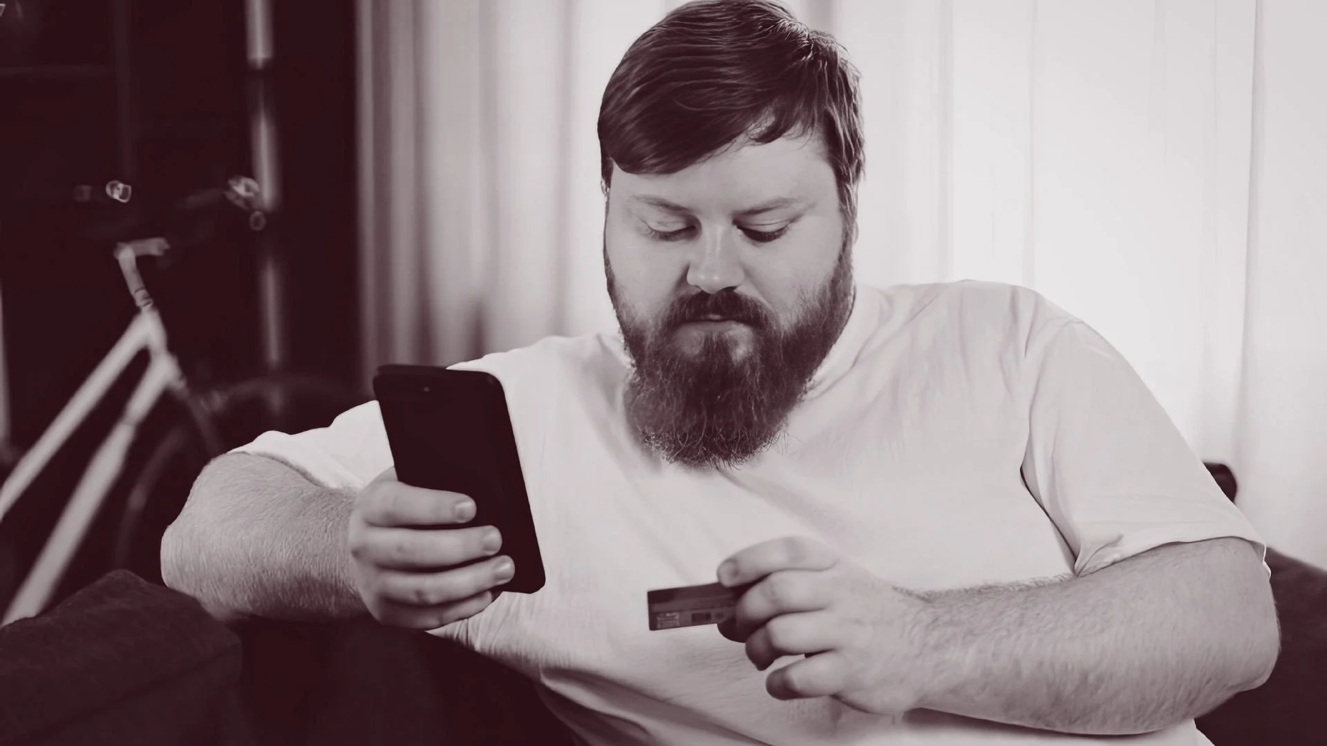 Man holding a credit card and looking at his phone, checking his bank balance at home