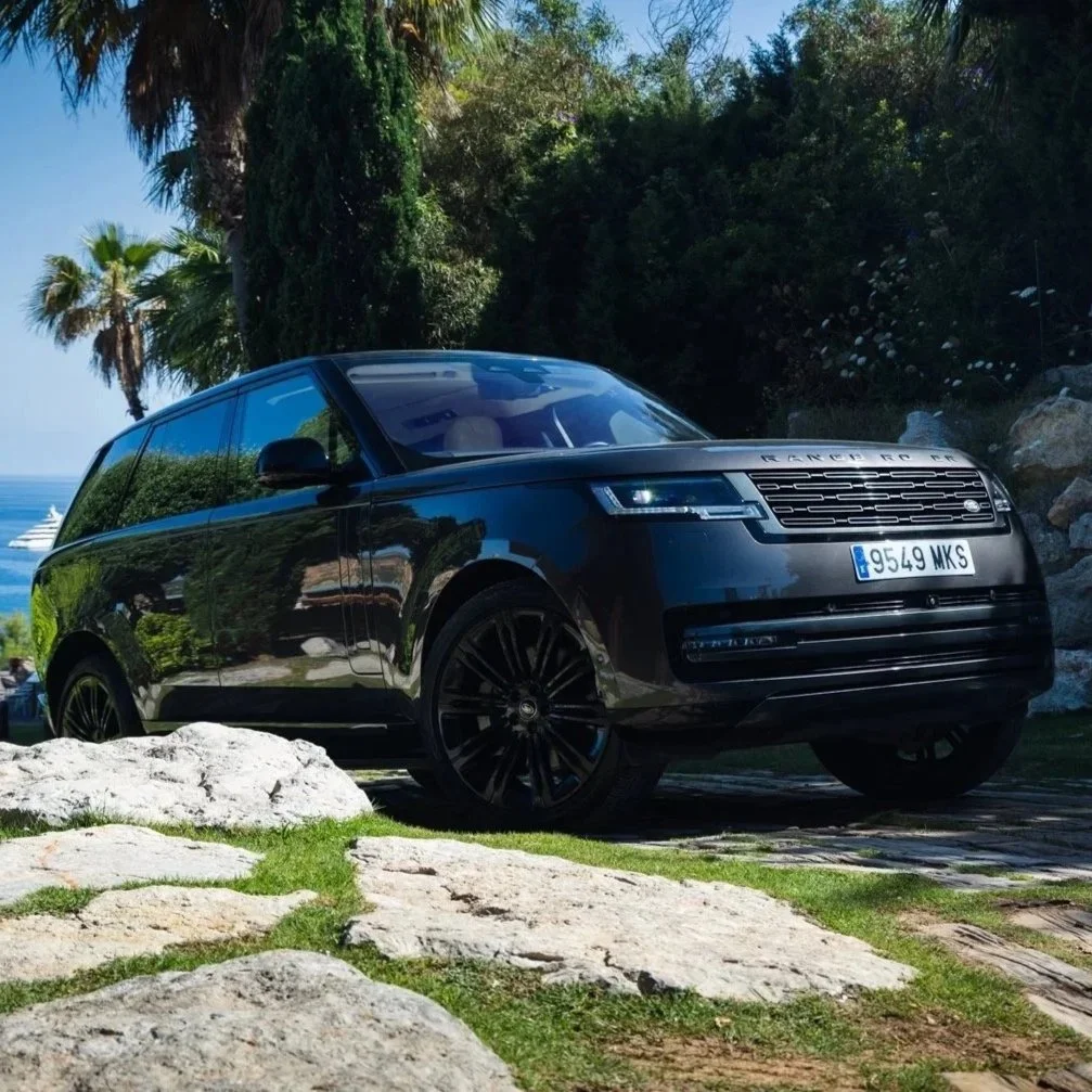 Black Range Rover SUV parked on a rocky, grassy area near palm trees and greenery, with the ocean and a yacht visible in the background.