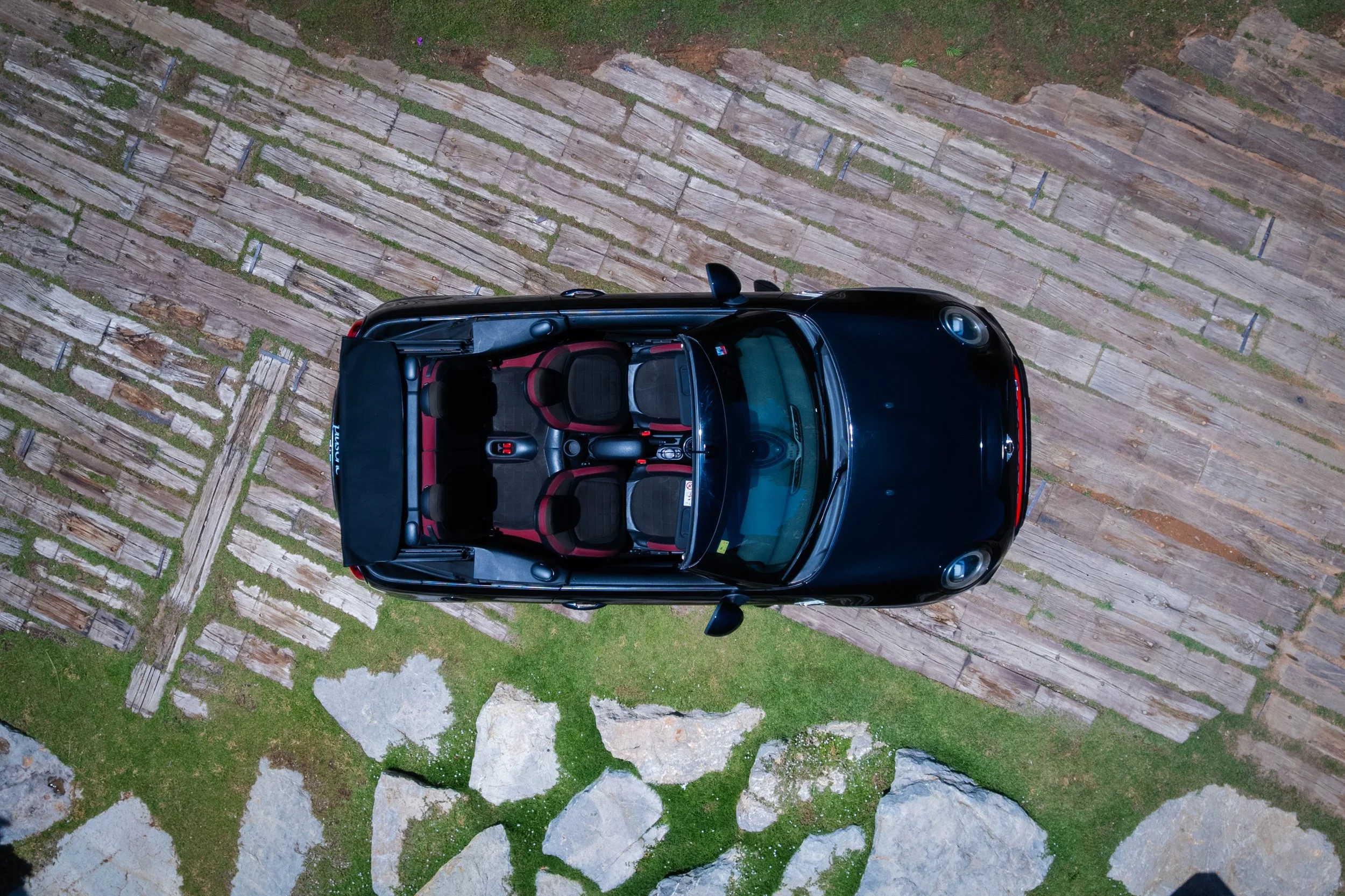 Top-down view of a black convertible car with red interior parked on a stone and grass pathway.
