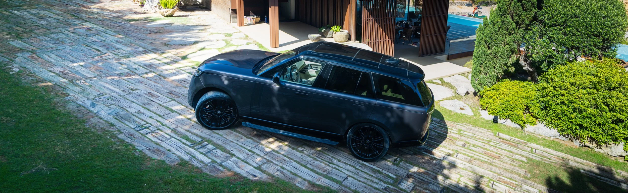 A black SUV parked on a stone driveway outside a modern house with a garden and a swimming pool.