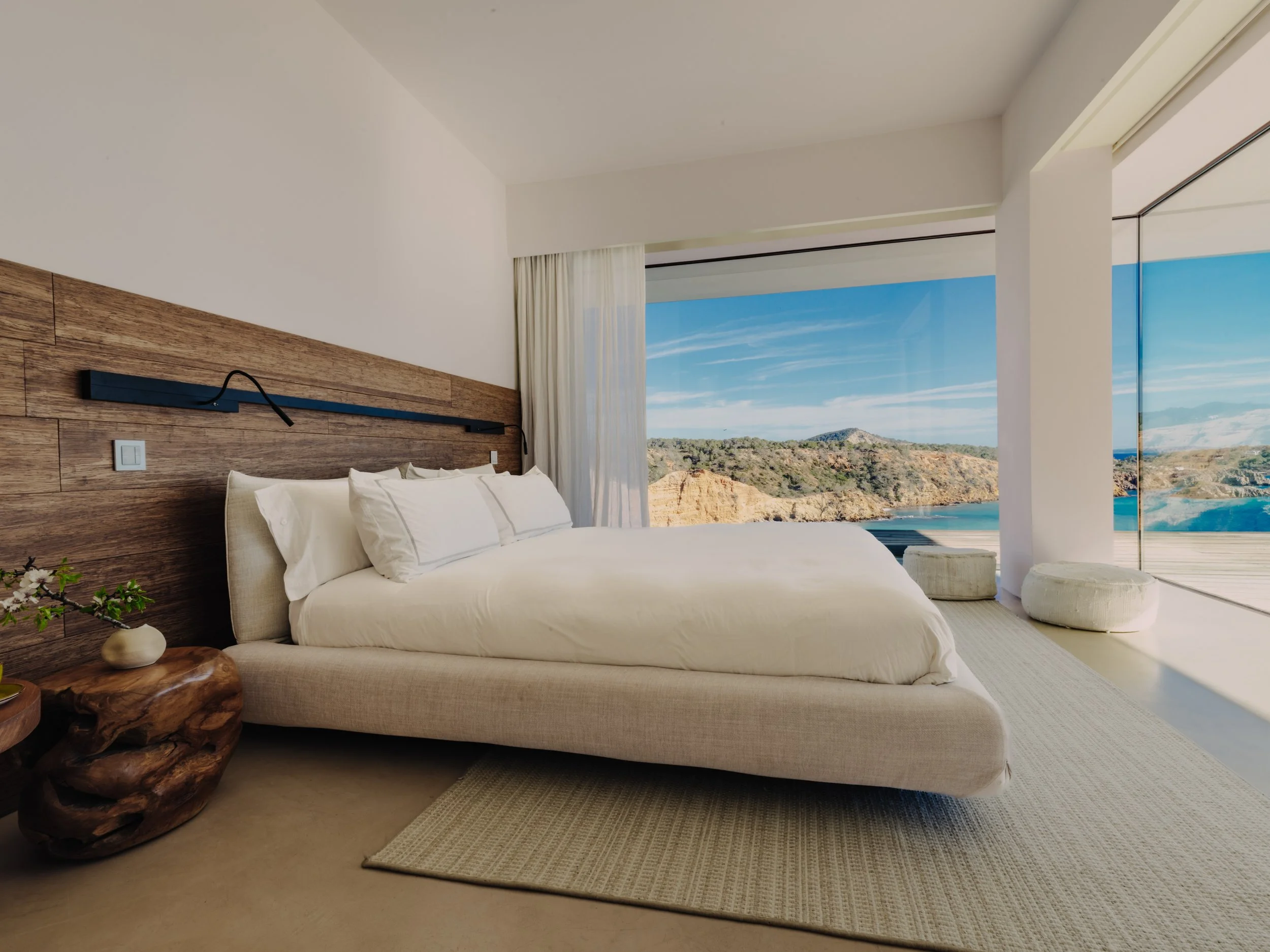 A bedroom with a large bed, wooden headboard, and view of scenic cliffs and blue sky through large windows.