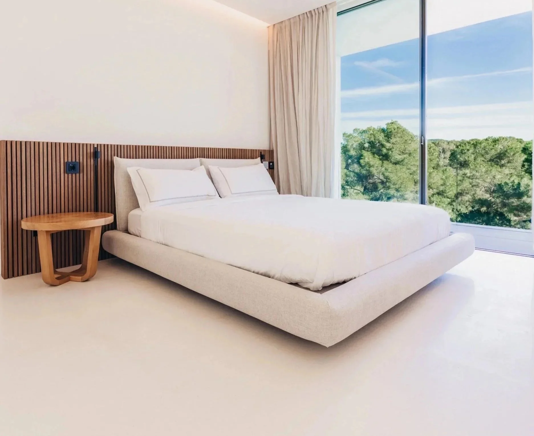 Modern hotel room with a large bed, white bedding, and a wooden headboard, next to a large glass window showing green trees and a blue sky with wispy clouds.