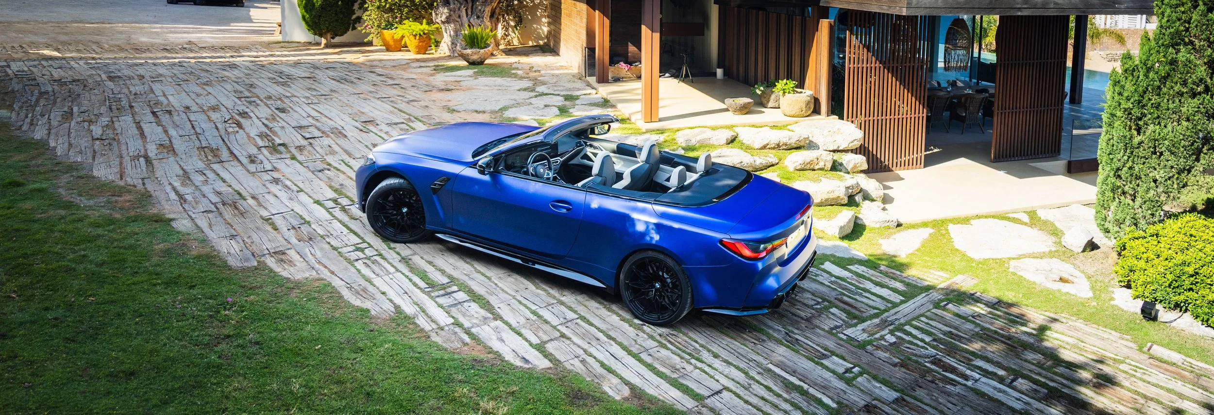 Blue convertible sports car parked on a stone driveway in front of a modern house with wooden and glass exterior, surrounded by greenery and potted plants.