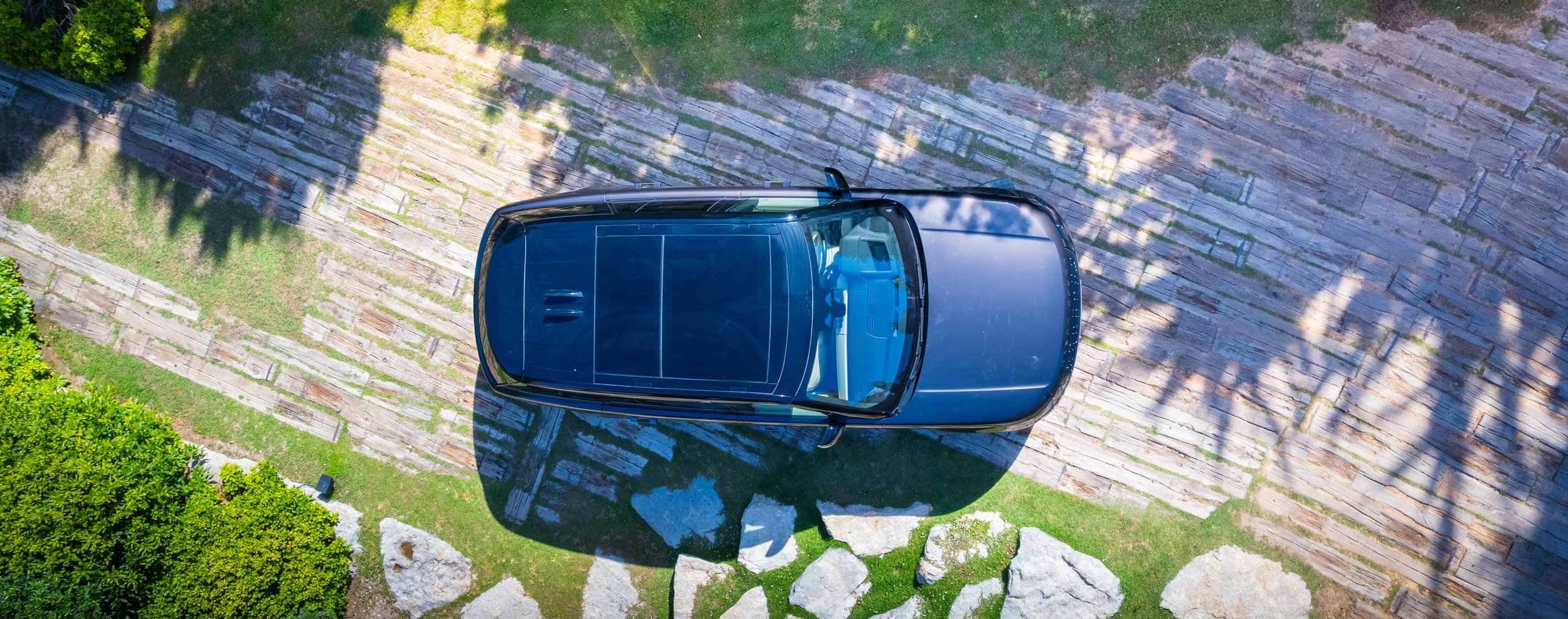 top-down aerial view of a black car parked on a stone driveway surrounded by greenery and rocks, with shadows of trees cast across the scene.