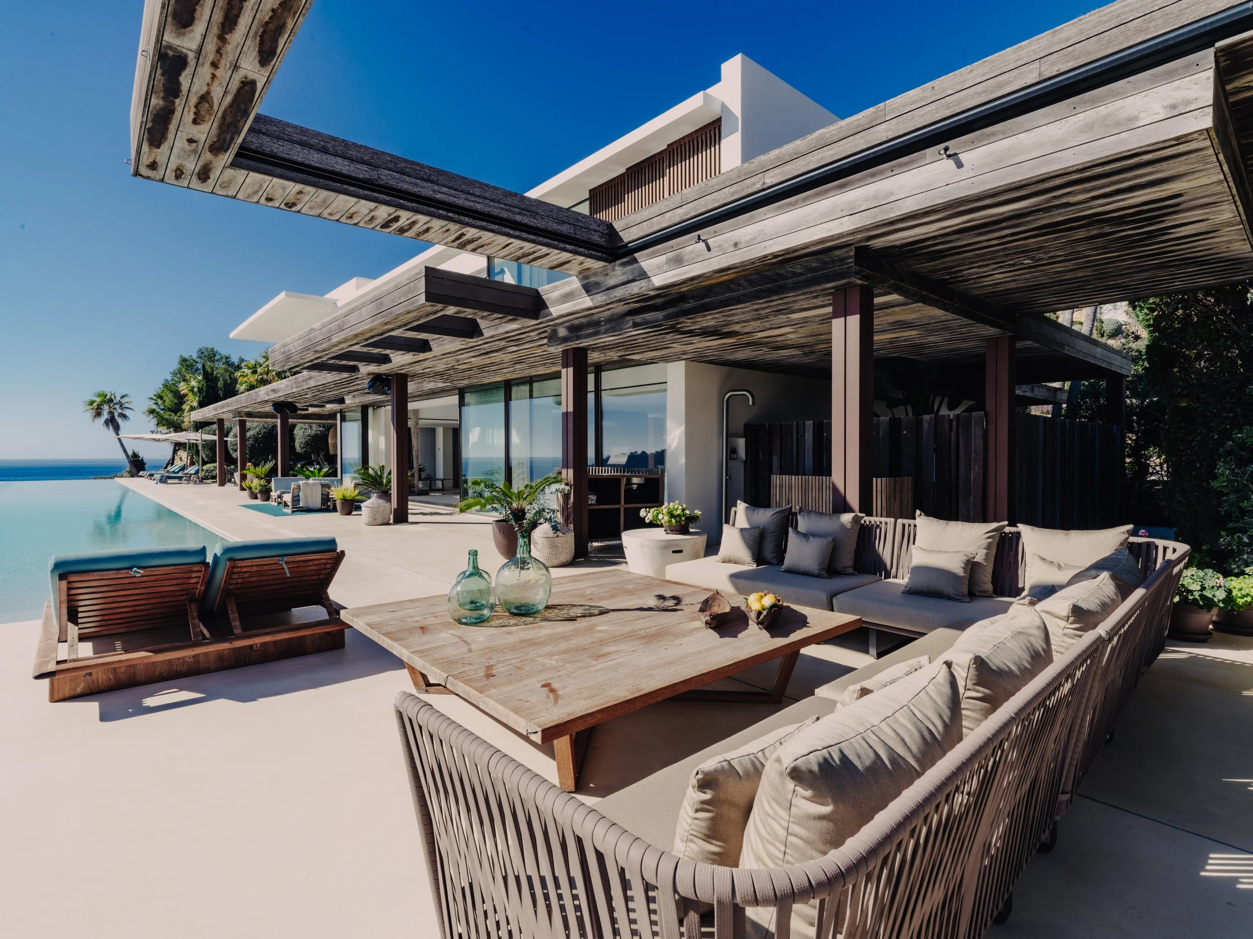 Modern house with an outdoor patio featuring a pool, cushioned lounge chairs, and a large wooden table with glass vases, under a wooden pergola, against a background of blue sky, palm trees, and ocean.