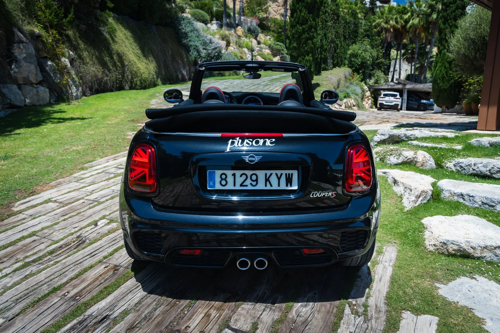 Rear view of a black Mini Cooper convertible with a personalized license plate, parked on a wooden pathway, with a garden and other cars in the background.