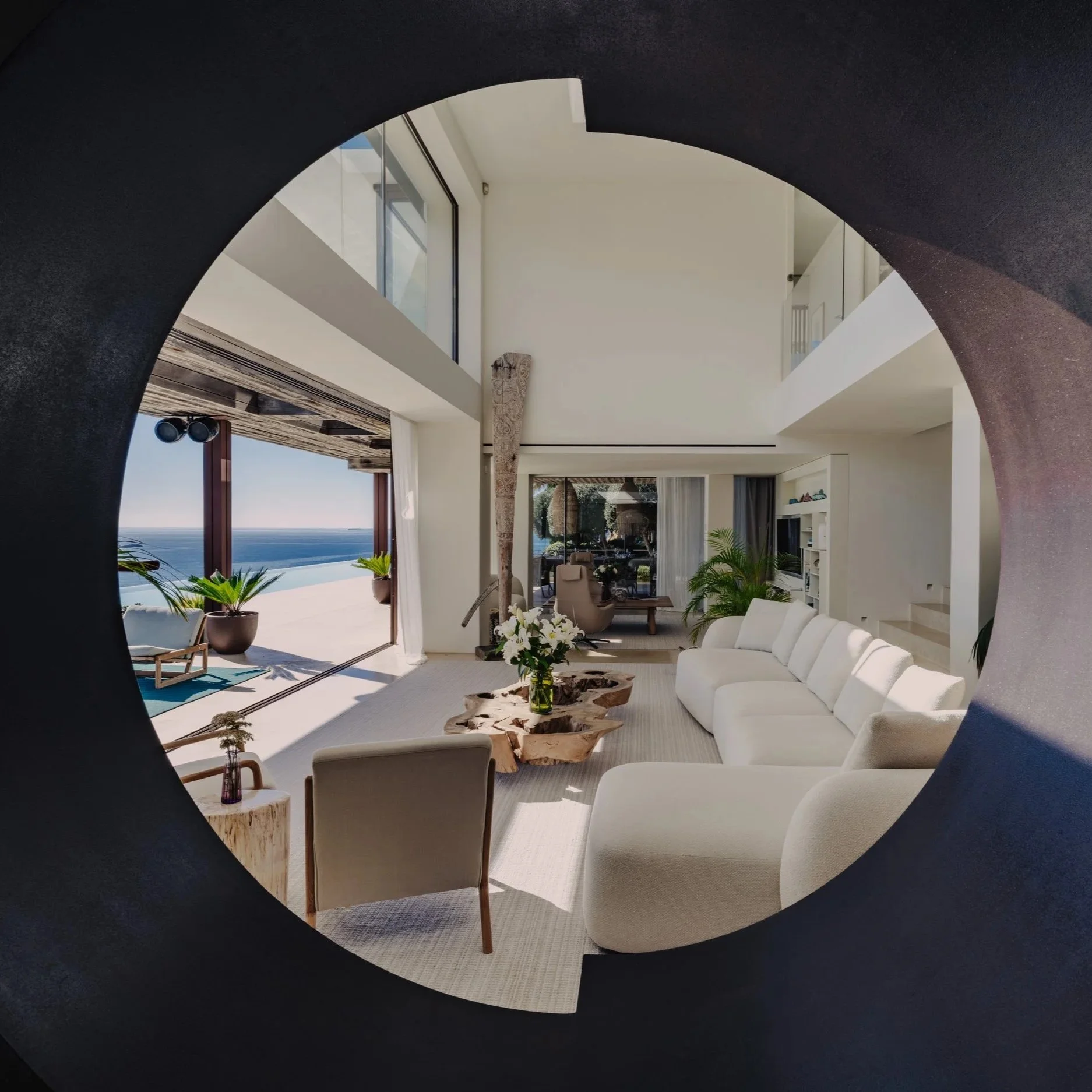 Modern living room with white sofas, a unique wooden coffee table, plants, and floor-to-ceiling windows overlooking the ocean.