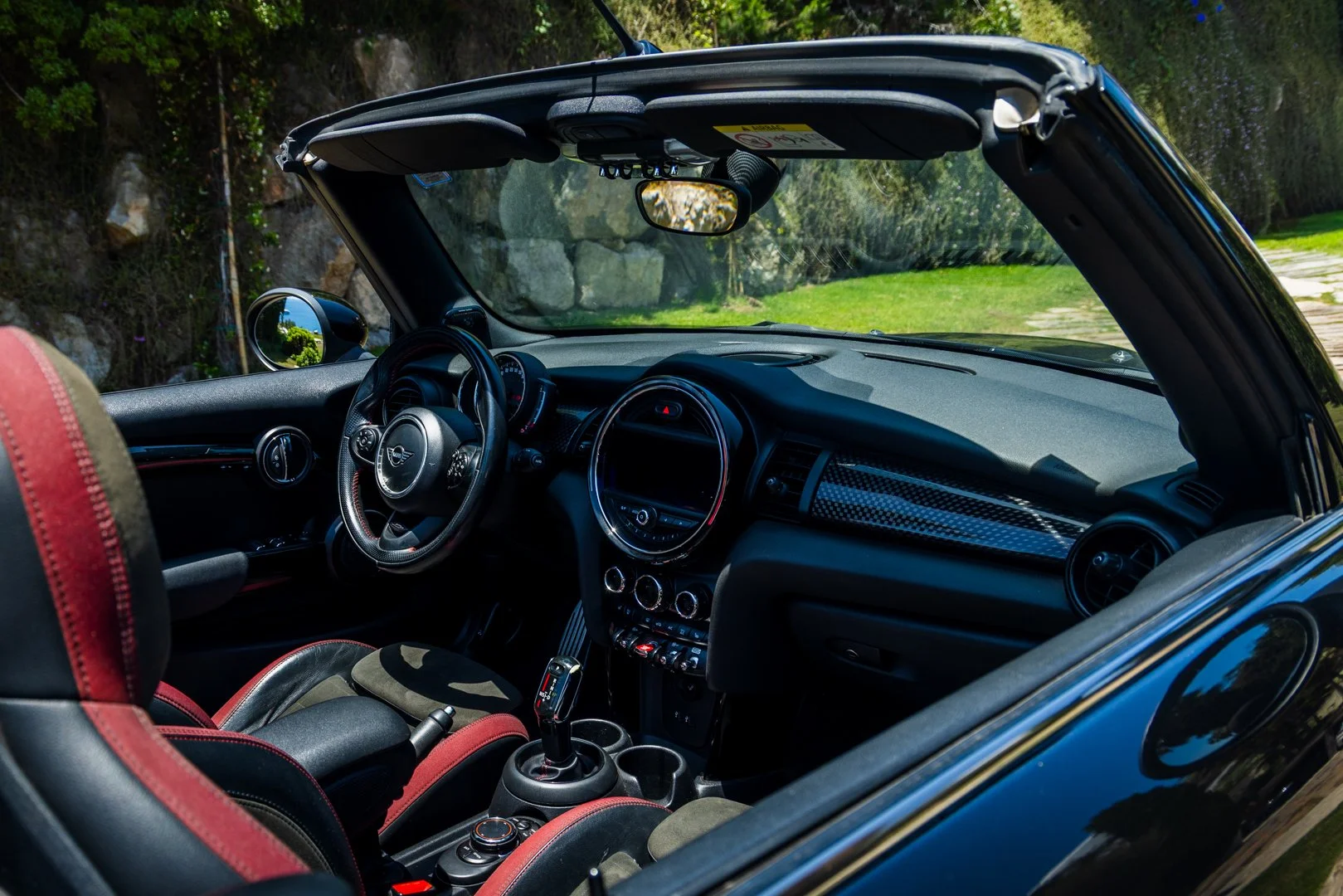 Inside view of a black Mini Cooper convertible with red accents, showing the dashboard, steering wheel, and seats, parked on a scenic road with greenery and rocks outside.