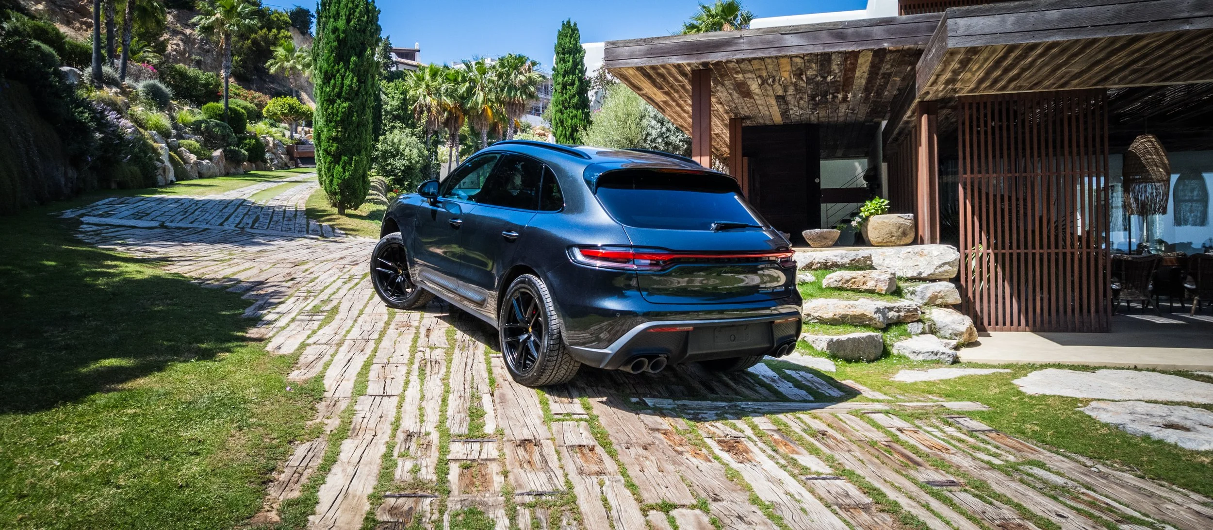 A black luxury SUV parked on a stone-paved driveway in front of a modern house with a wooden pergola, surrounded by greenery and palm trees.