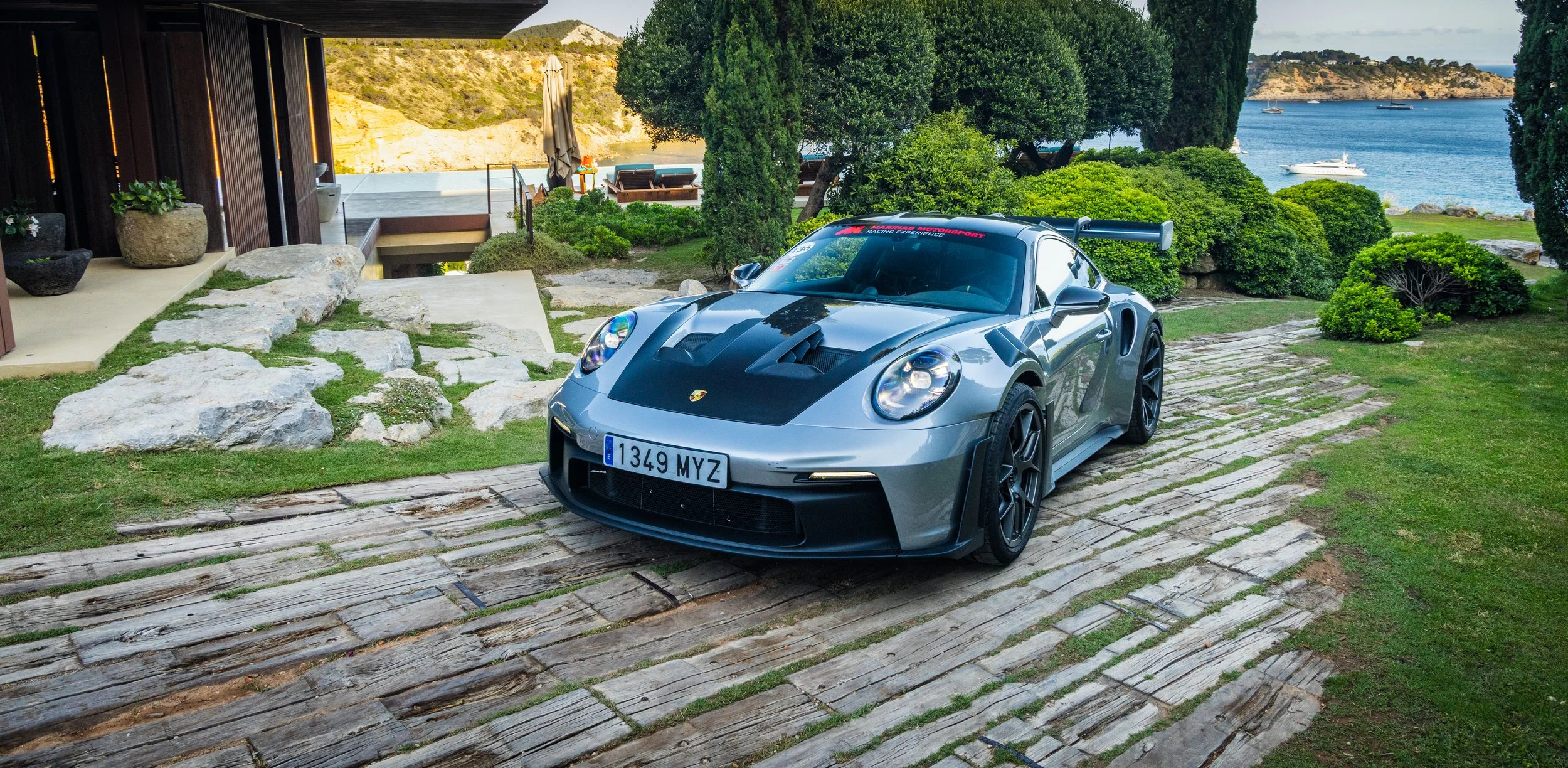 A silver Porsche sports car parked on a stone driveway near a modern house with a view of the ocean and boats in the background.