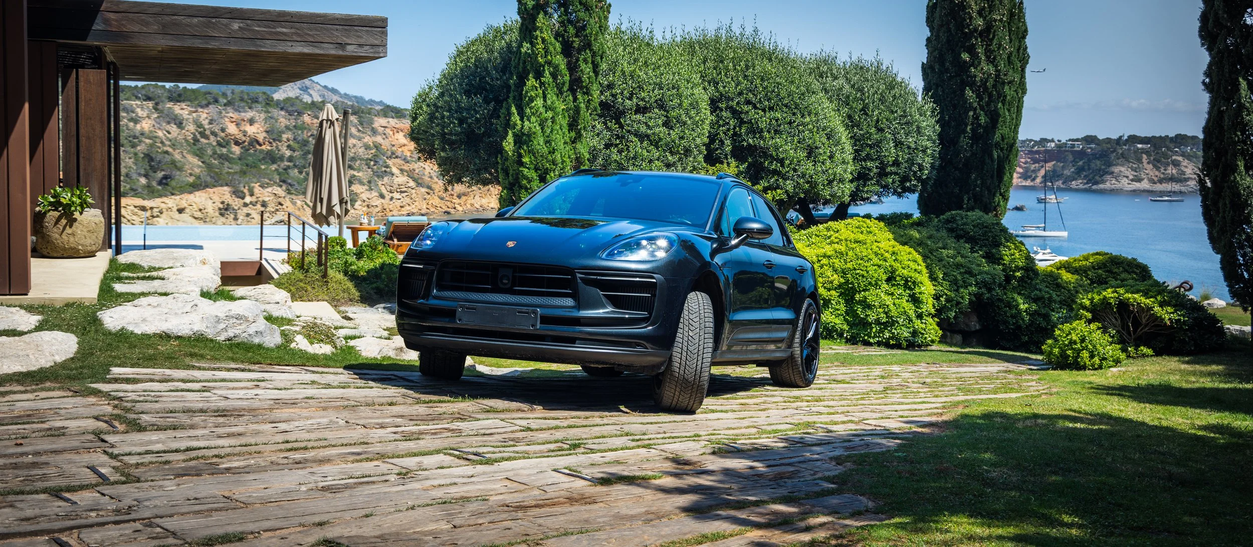 A black Porsche SUV parked on a stone driveway in front of a house with lush green trees and a view of a bay with sailboats and distant hills.