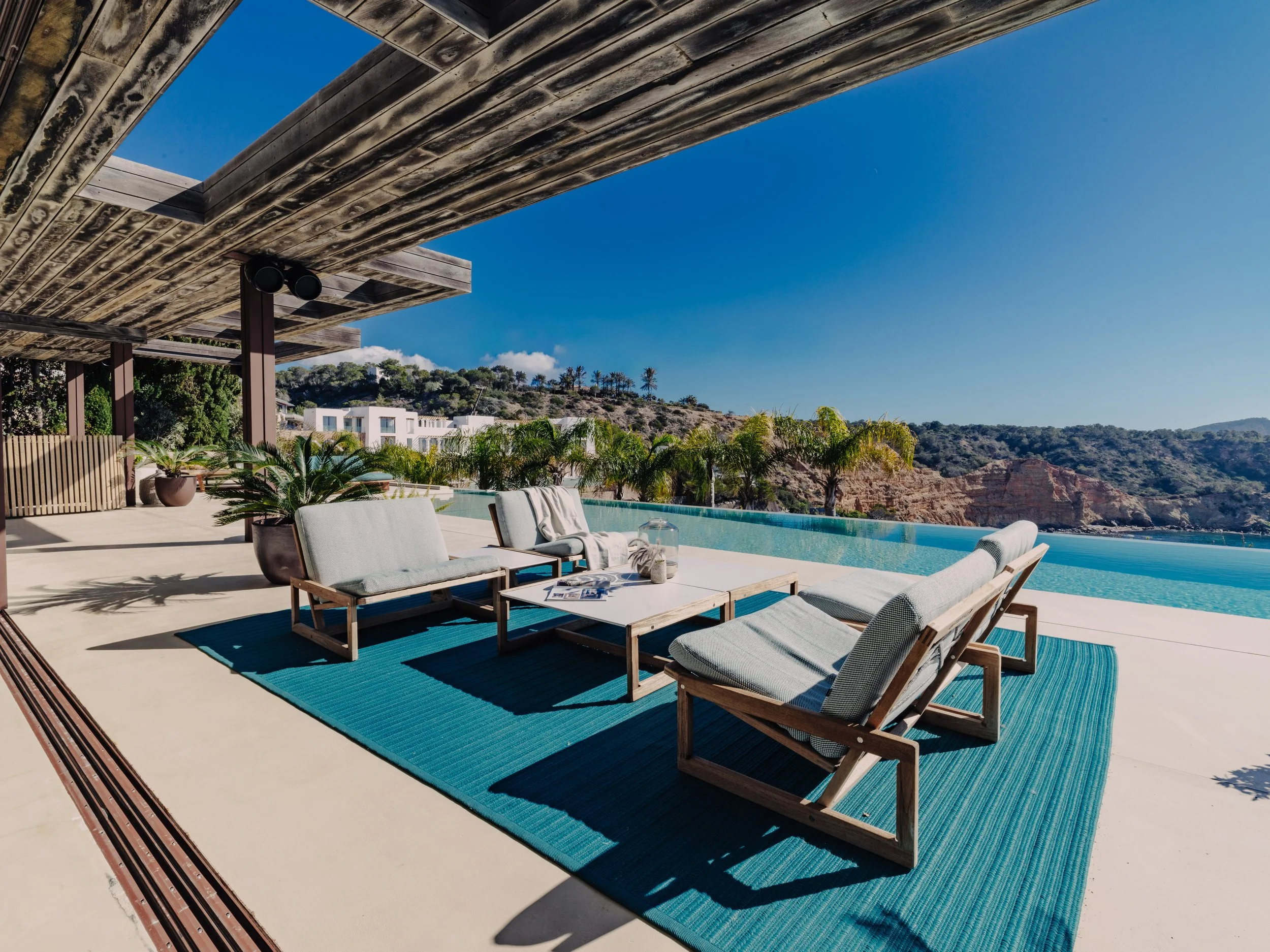 Outdoor patio area with wooden furniture, potted plants, a swimming pool, and a view of hills with trees and white buildings under a clear blue sky.
