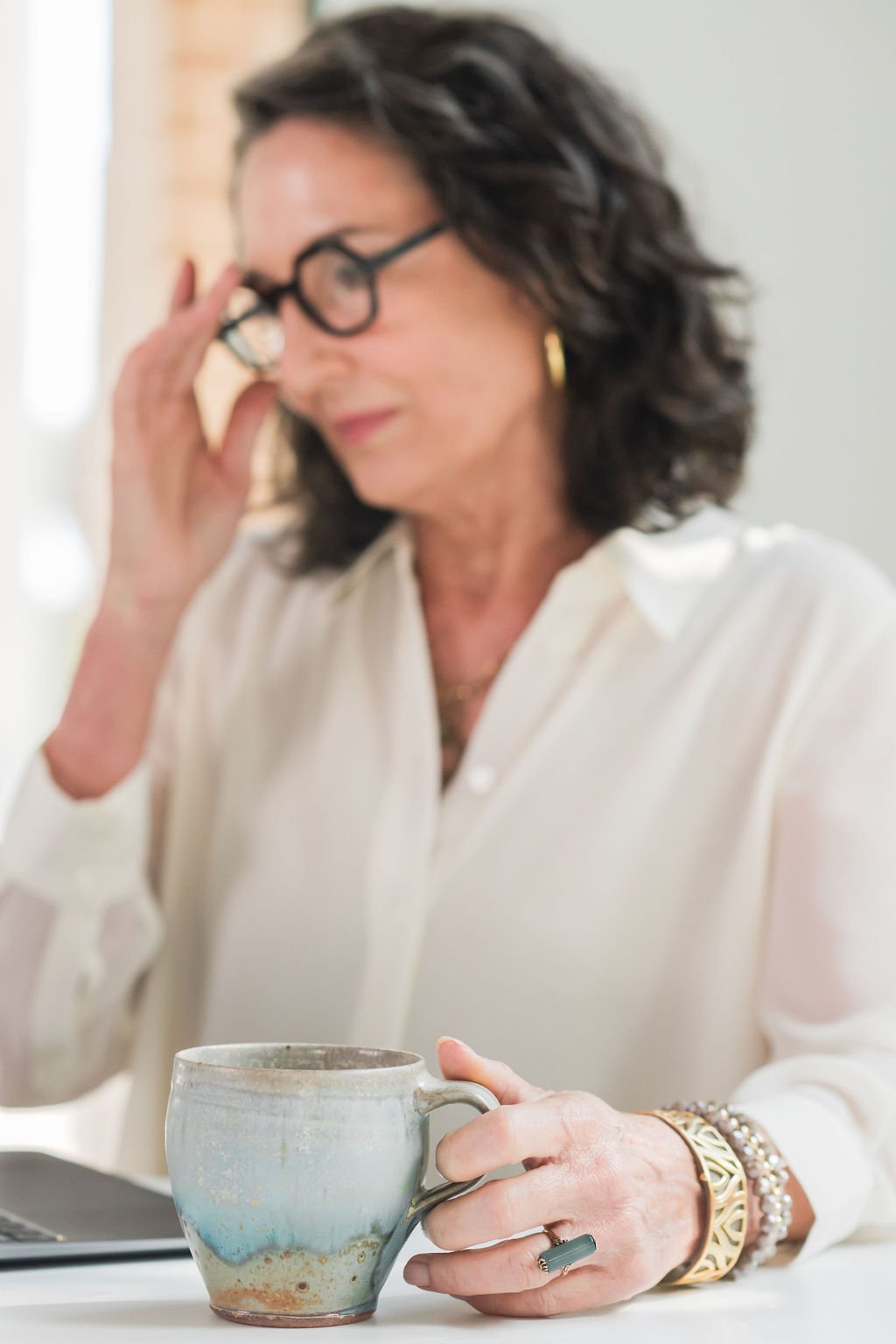 A woman with curly dark hair, wearing glasses and a white blouse, is sitting at a table holding a ceramic mug in her right hand. She appears to be thinking or concentrating, with her left hand touching her forehead. She is wearing jewelry including bracelets and a ring.