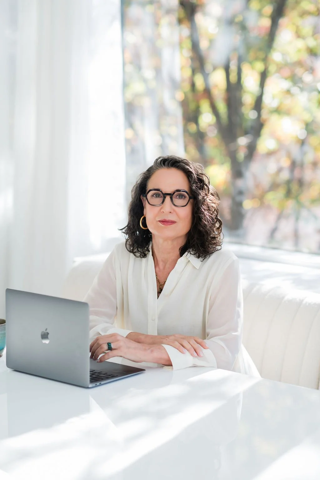 Debra Benfield, a woman with curly dark hair and glasses sitting at a white table with a silver laptop in front of her, in a bright room with large windows and autumn trees outside.